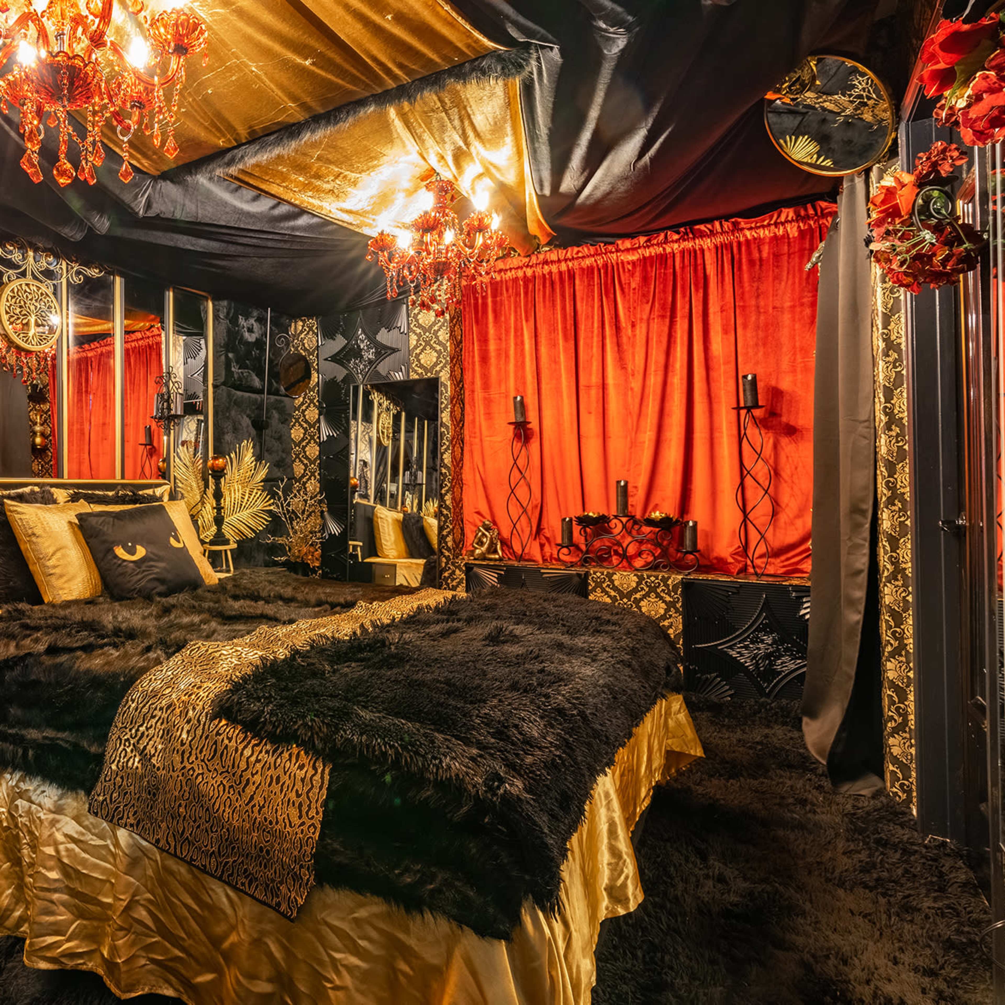 A luxurious bedroom decorated with black and gold accents, featuring a large bed covered with fur and surrounded by ornate mirrors and red drapes.