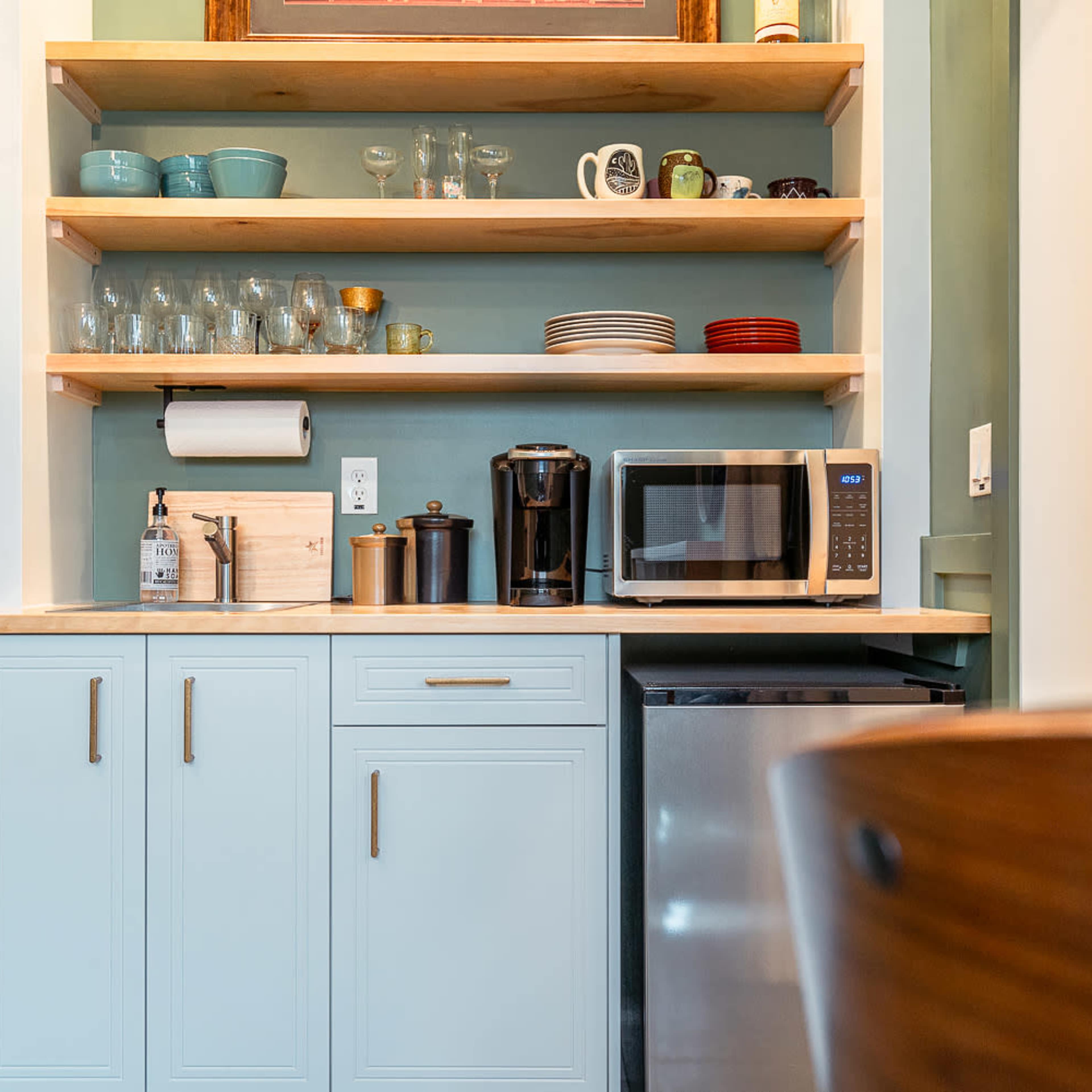 A kitchen area with open shelving displaying glassware and dishes, a microwave, a coffee maker, and a refrigerator beneath a counter.
