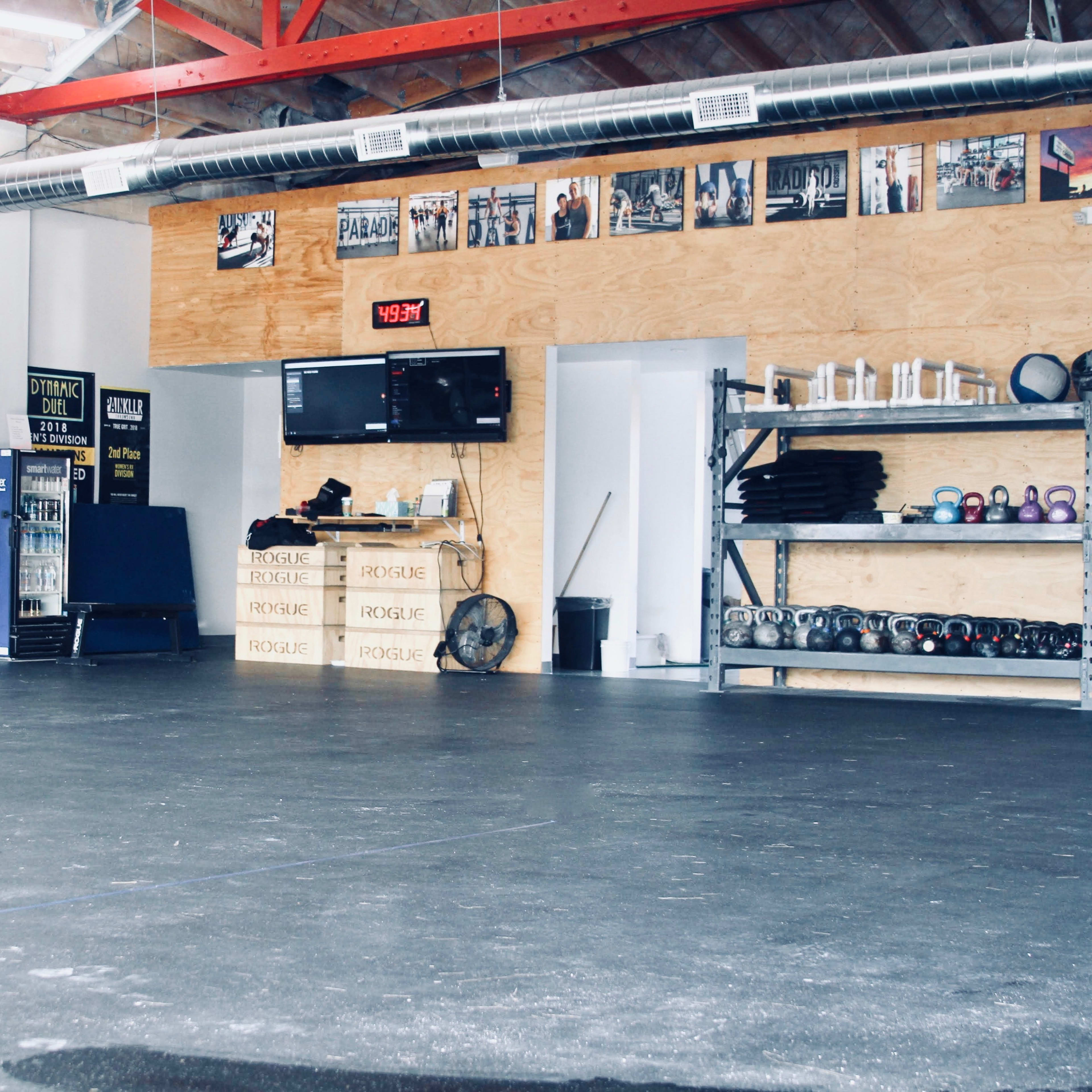 A spacious gym interior with wooden walls, workout equipment on shelves, a digital clock, and fitness posters on the walls.