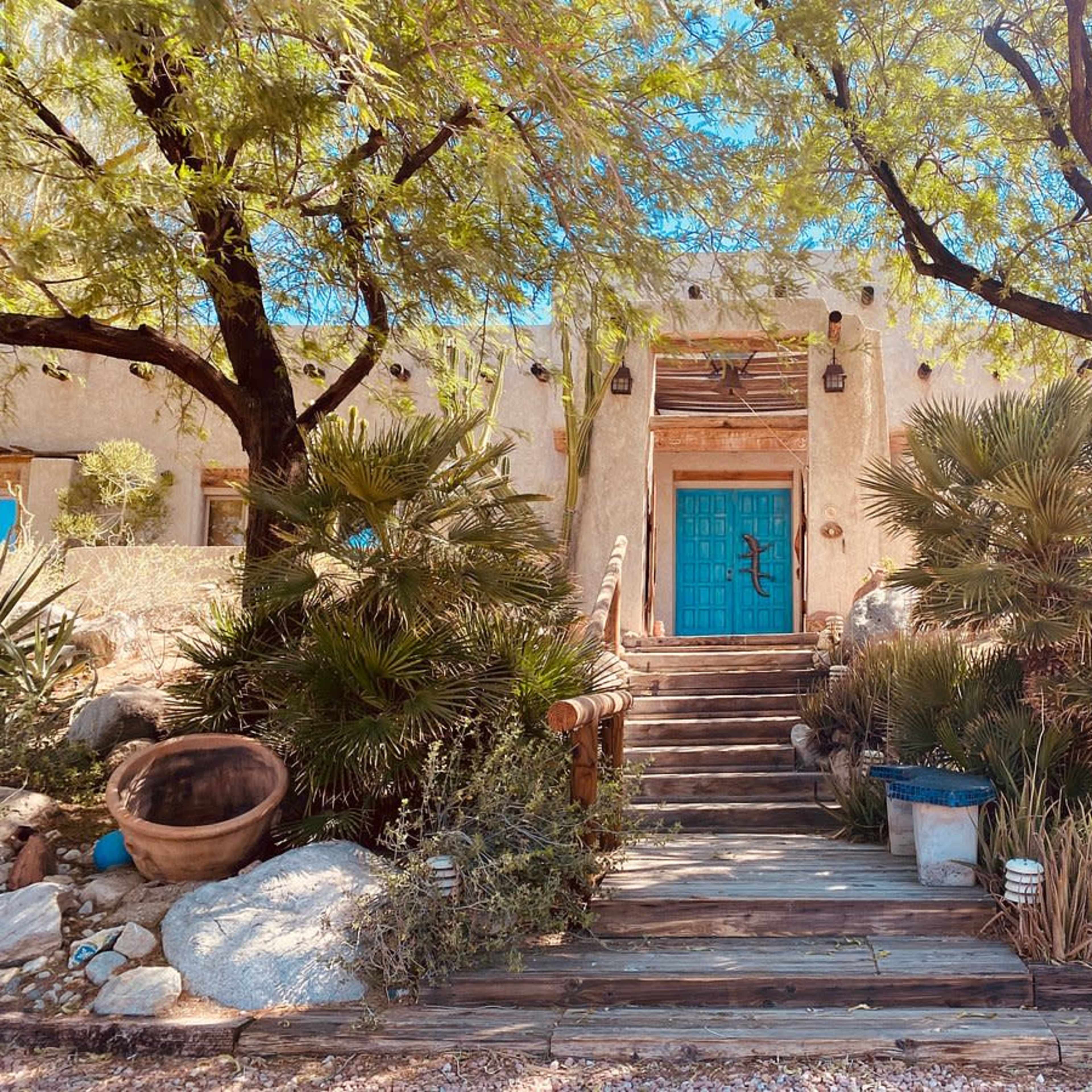 A staircase leads to a house with a blue door, surrounded by desert vegetation and trees.