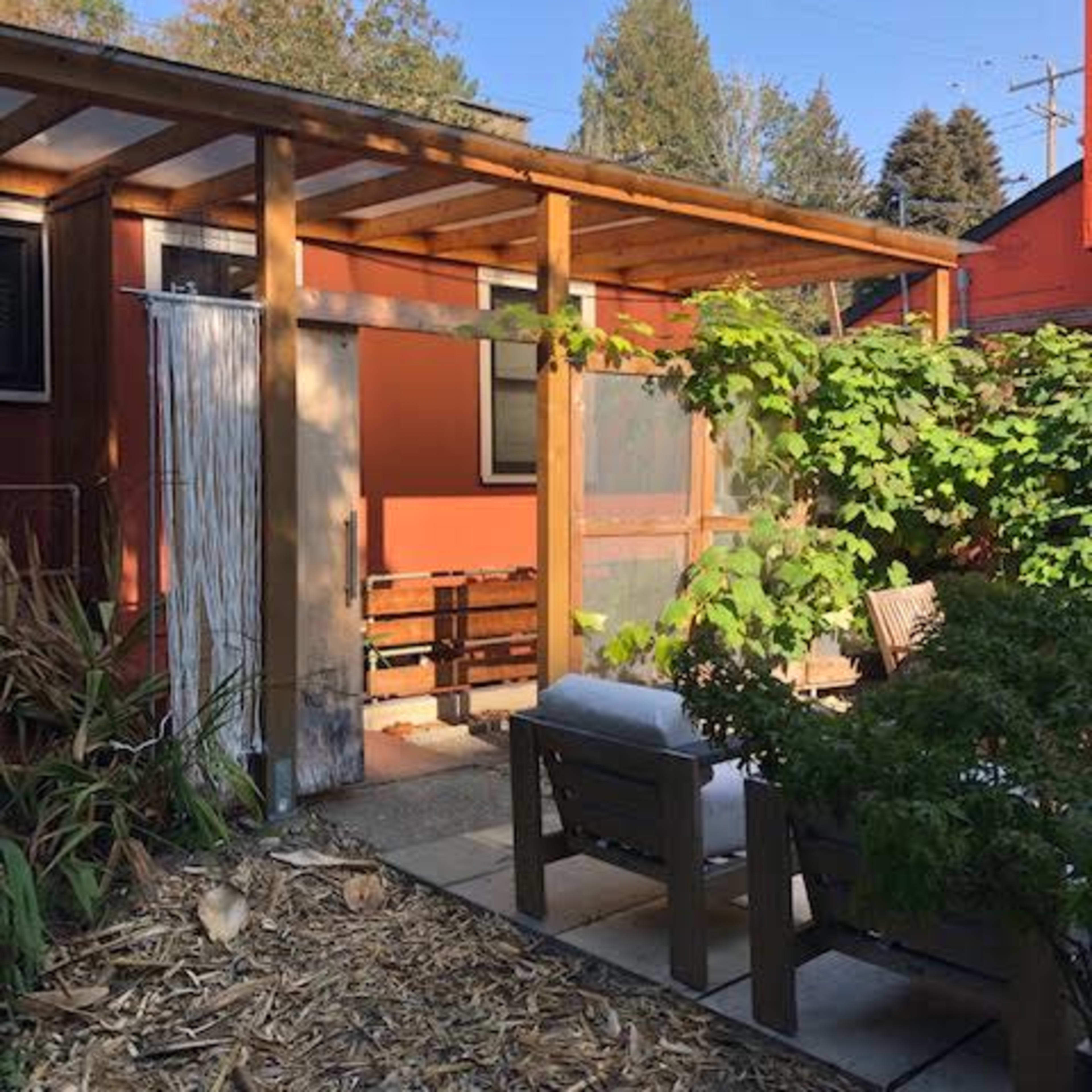 A backyard patio area with wooden furniture, surrounded by greenery and a small trellis, leading to a brightly colored house.