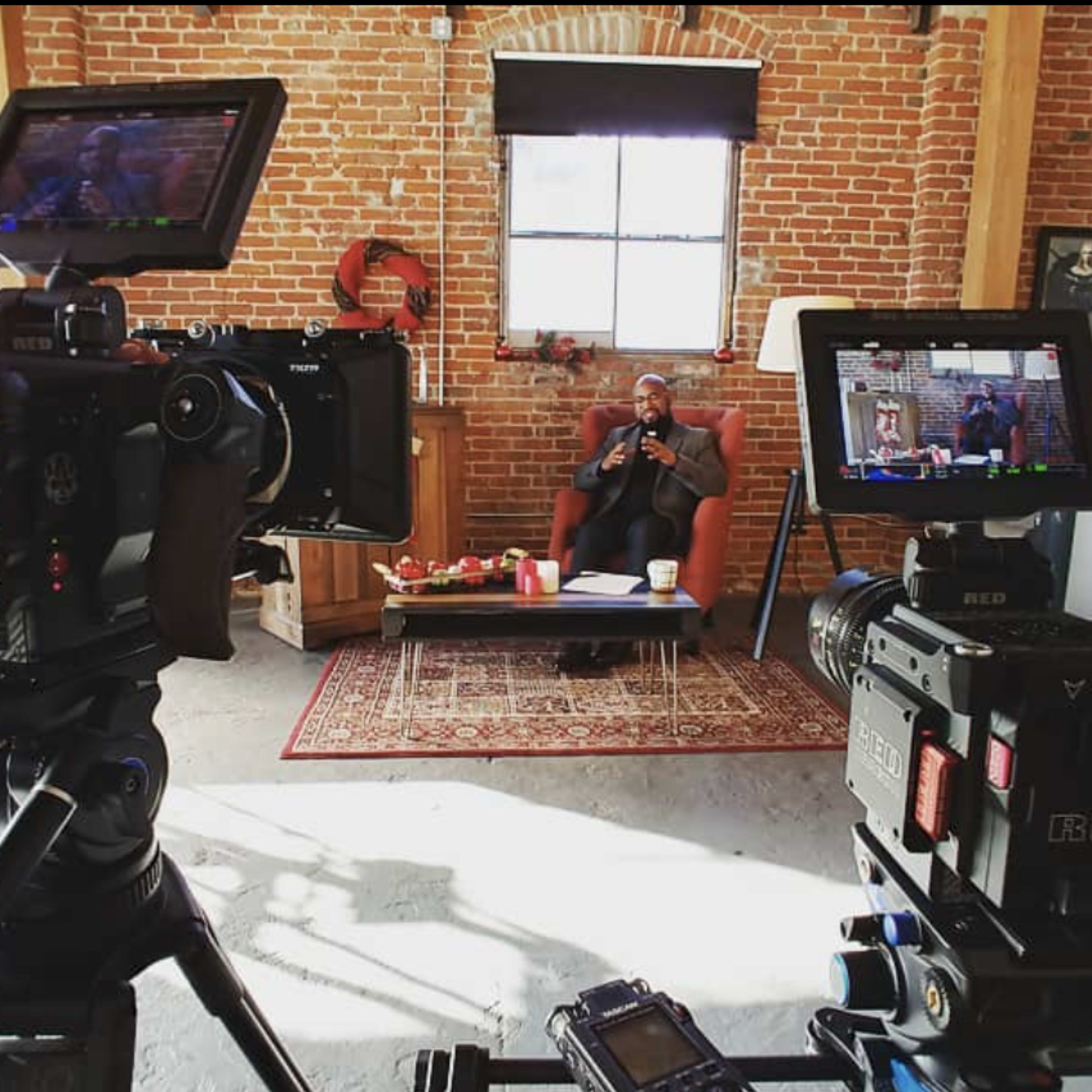 Two cameras positioned in the foreground, focusing on a person sitting in a studio space with brick walls, holding a device, and surrounded by a small table and decor.