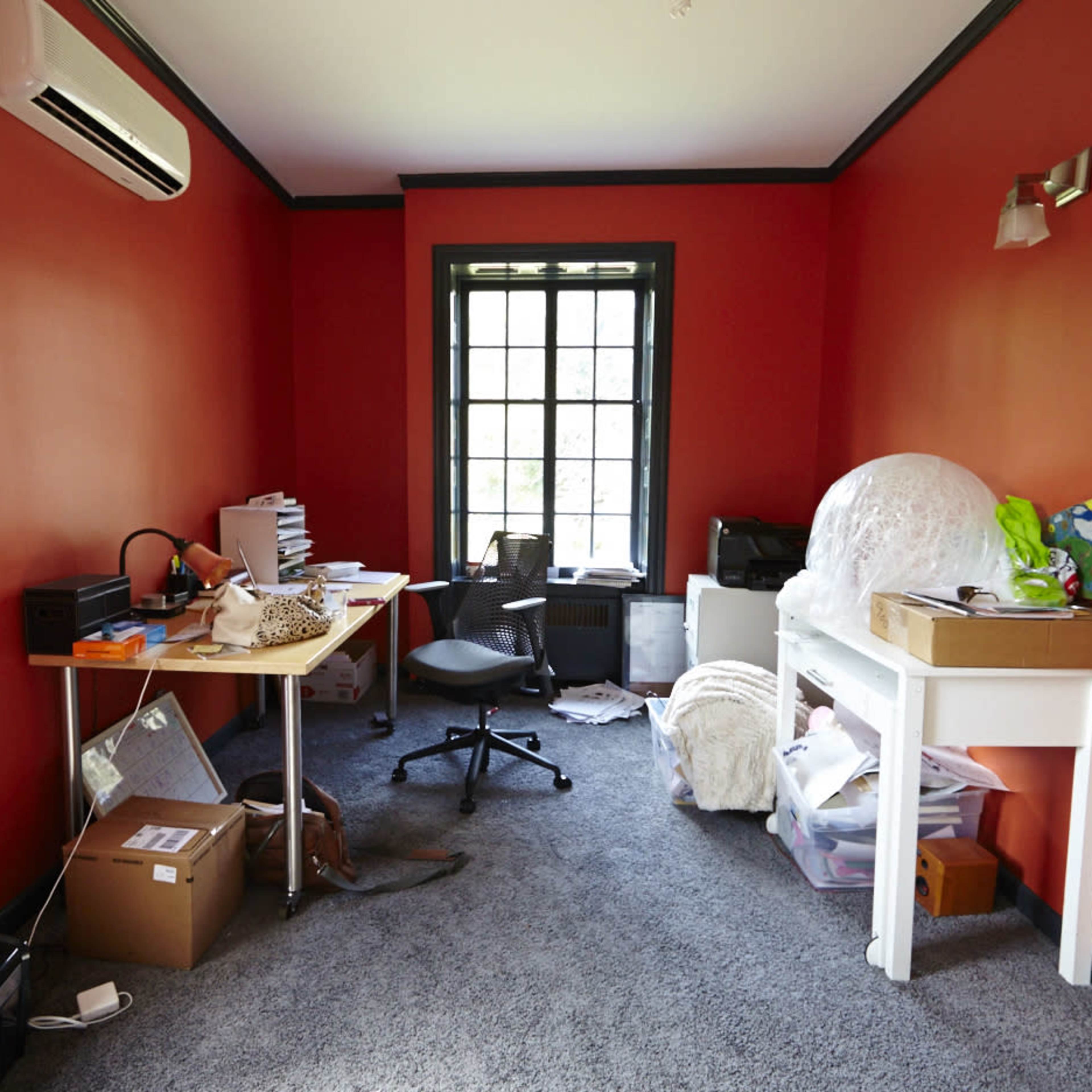 A room with red walls contains a desk, an office chair, piles of clutter, and a window with black trim.