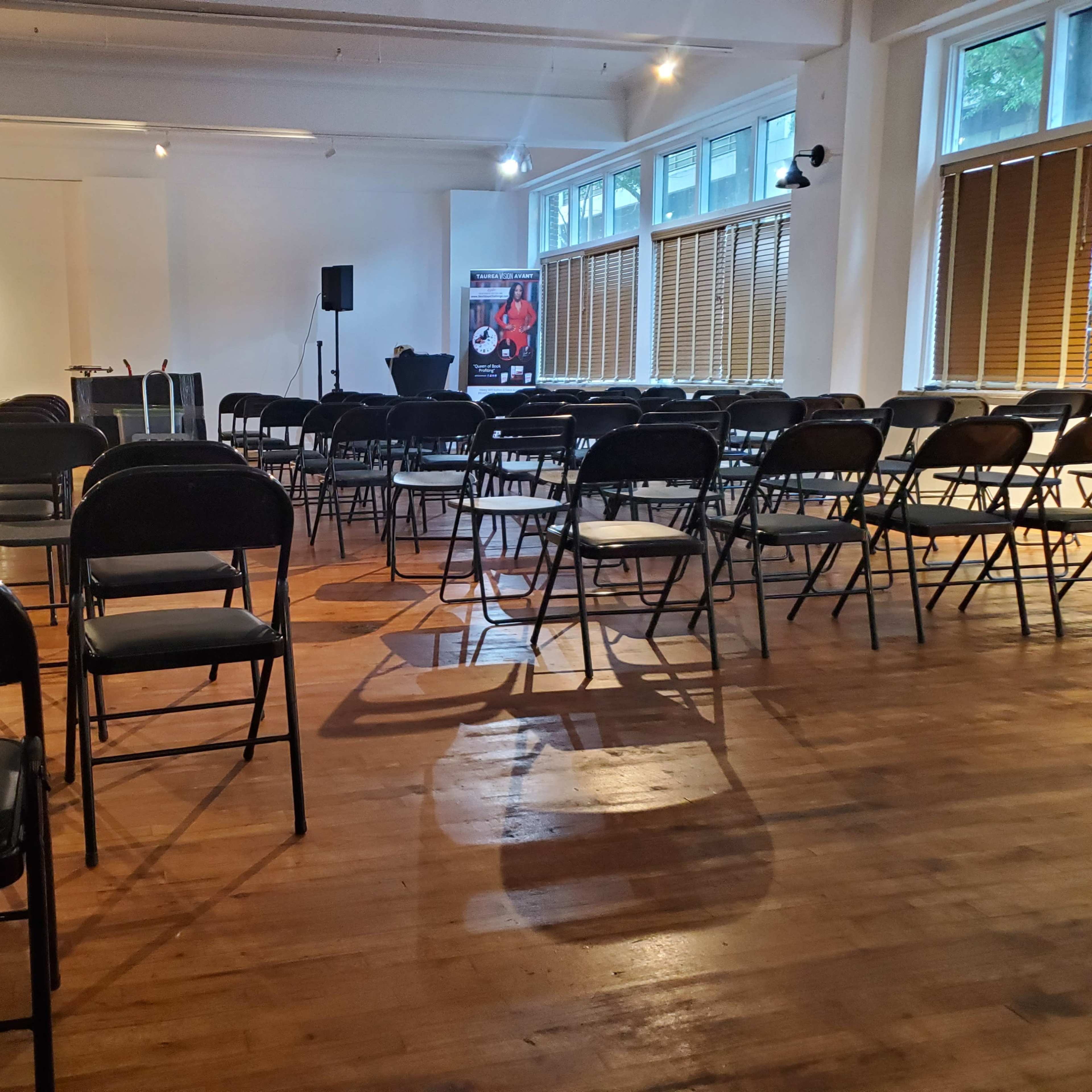 A spacious room with rows of black folding chairs arranged on a wooden floor, and a person walking towards the front where equipment is set up.