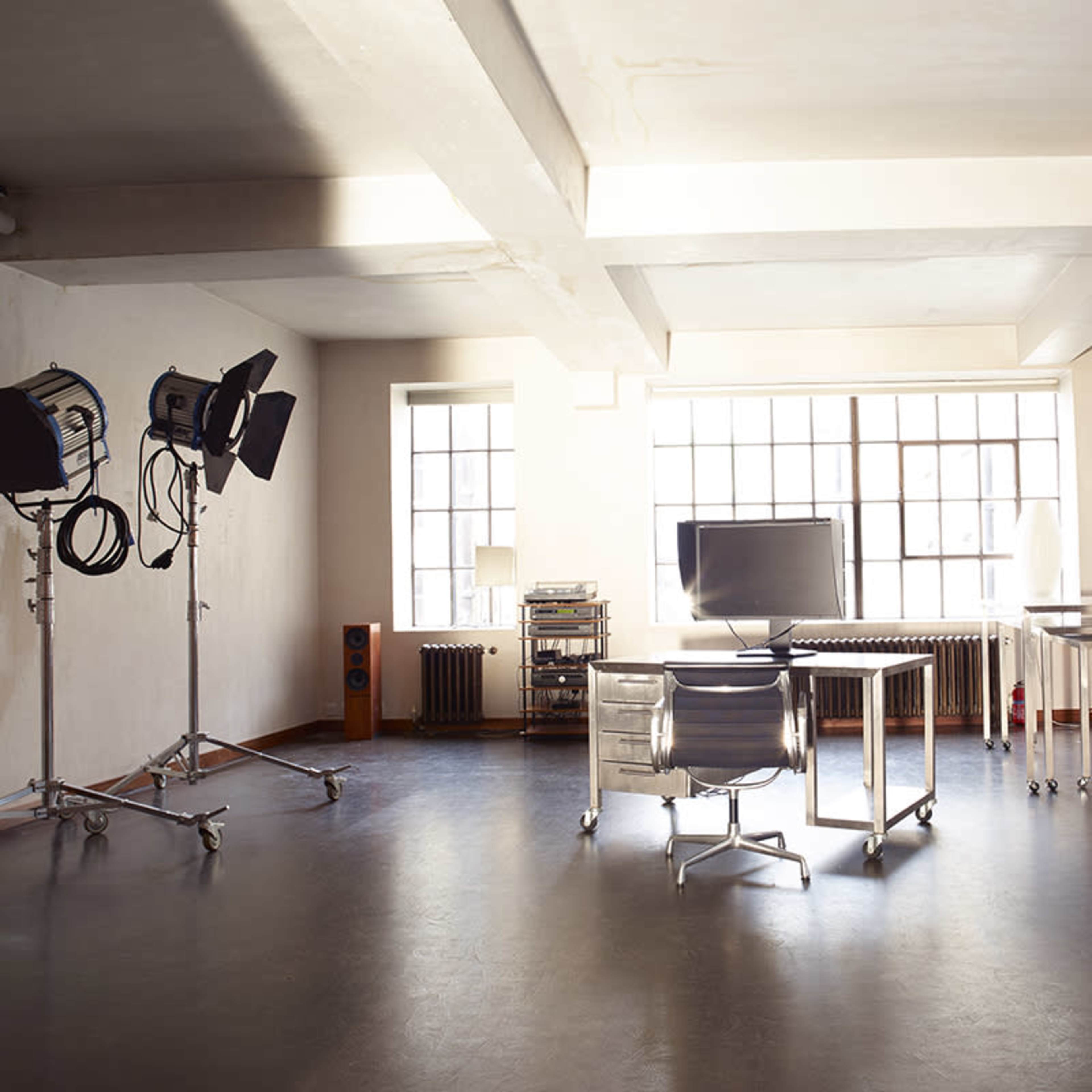 A spacious, minimalist studio with a desk, a computer monitor, and multiple lighting setups beside large windows.