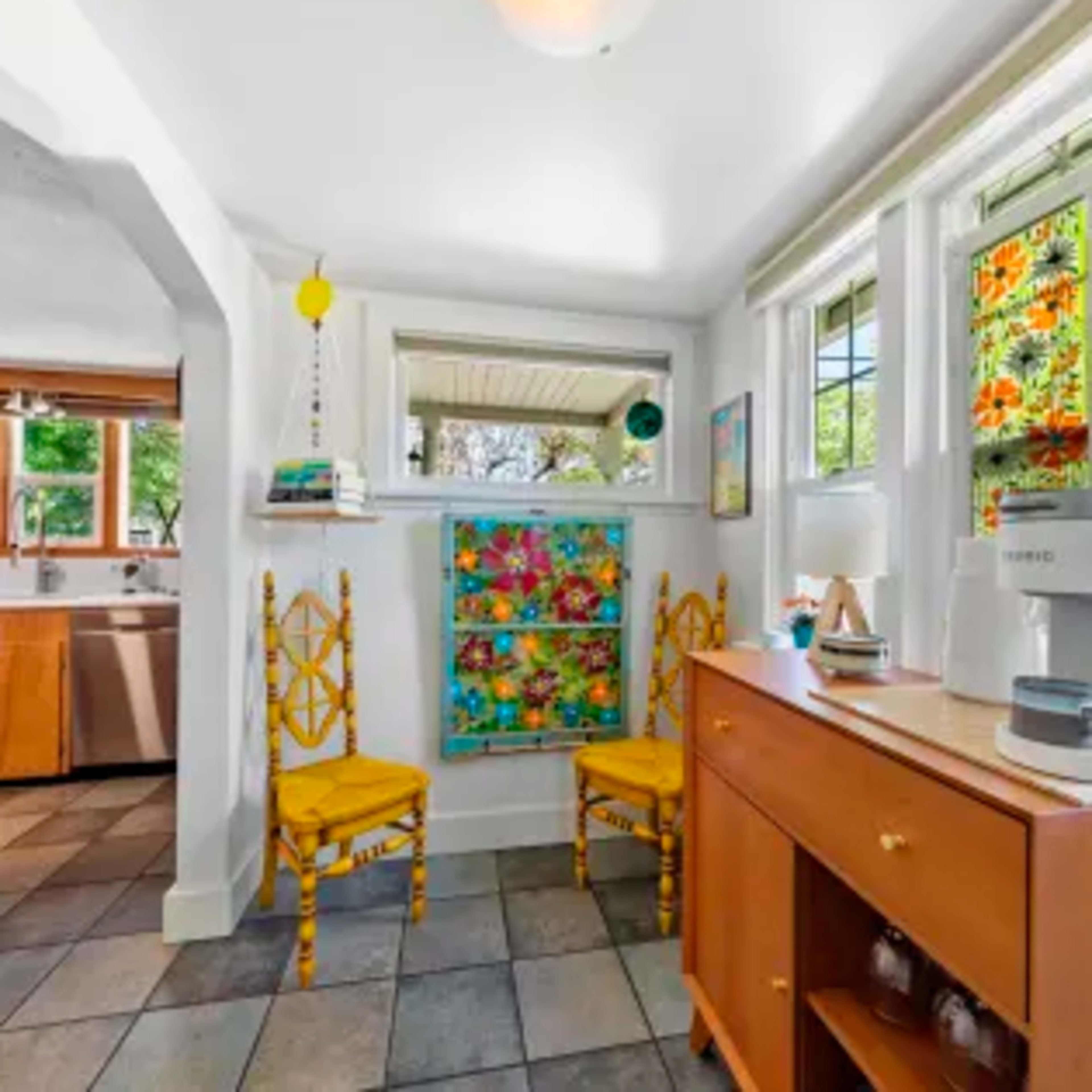 A brightly lit kitchen corner with two yellow chairs, a sideboard, and colorful stained glass windows.