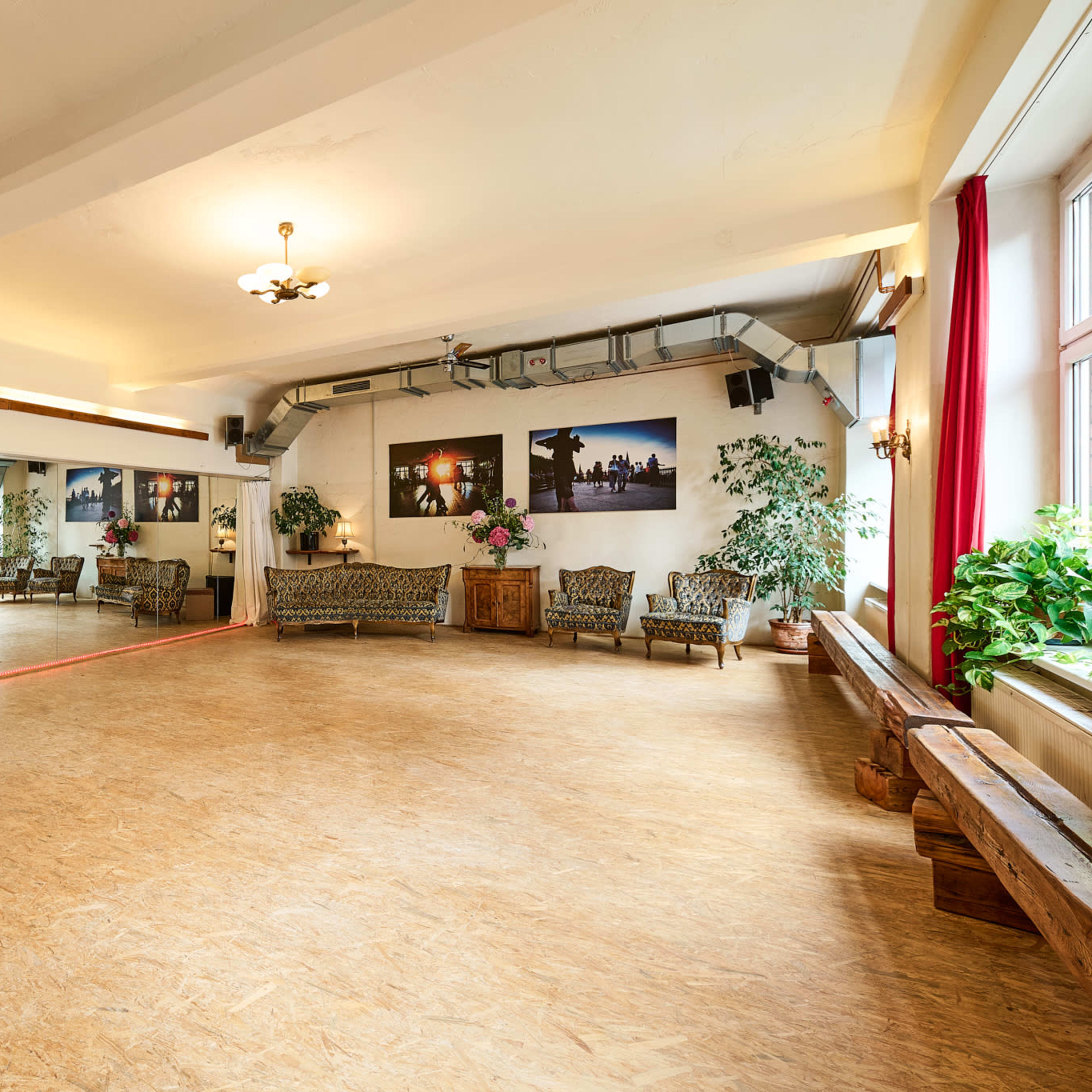 A spacious dance studio features a mirrored wall, wooden flooring, and decorative plants, with seating along the edges and large windows letting in natural light.