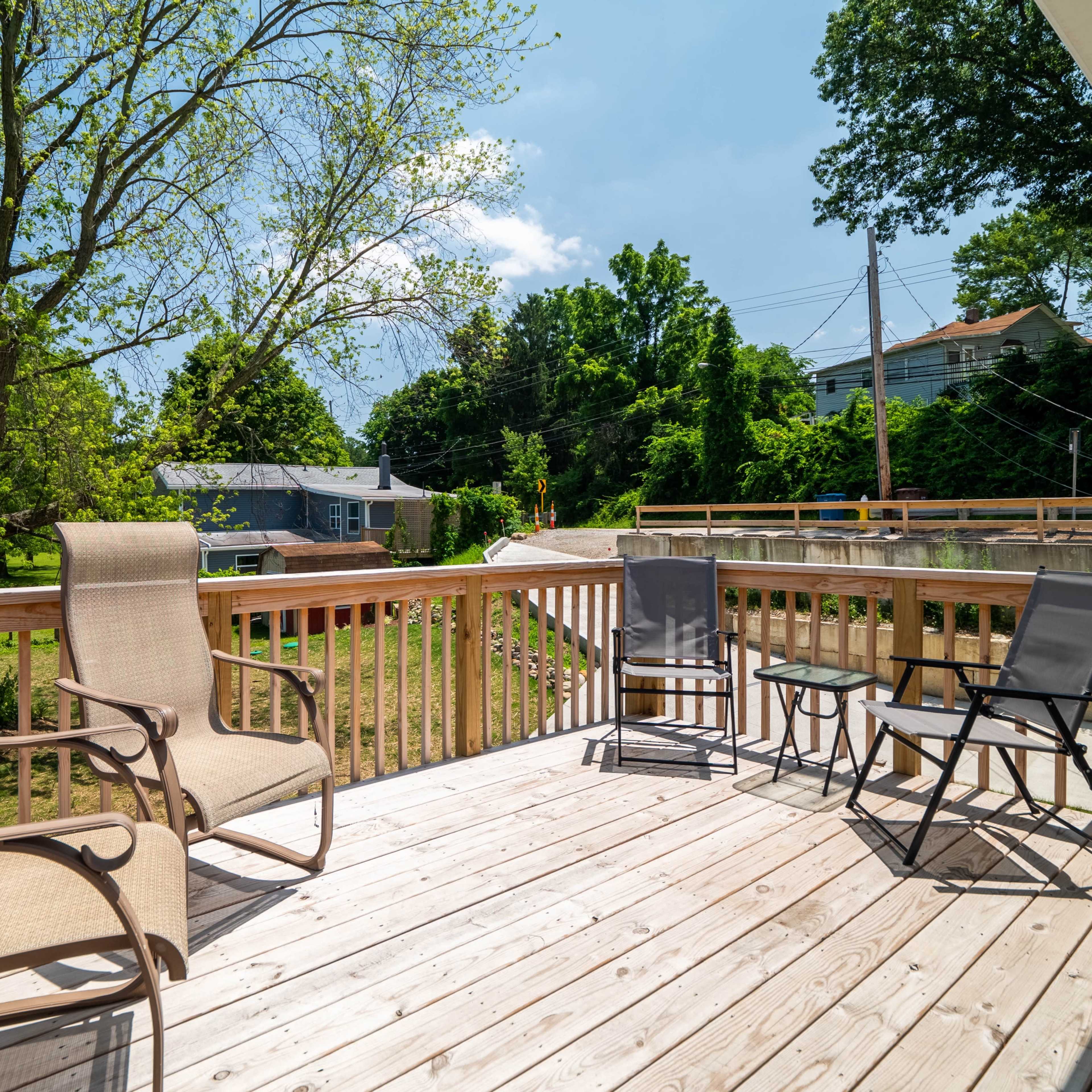 A wooden deck furnished with four chairs overlooking a green, tree-lined area and a road.