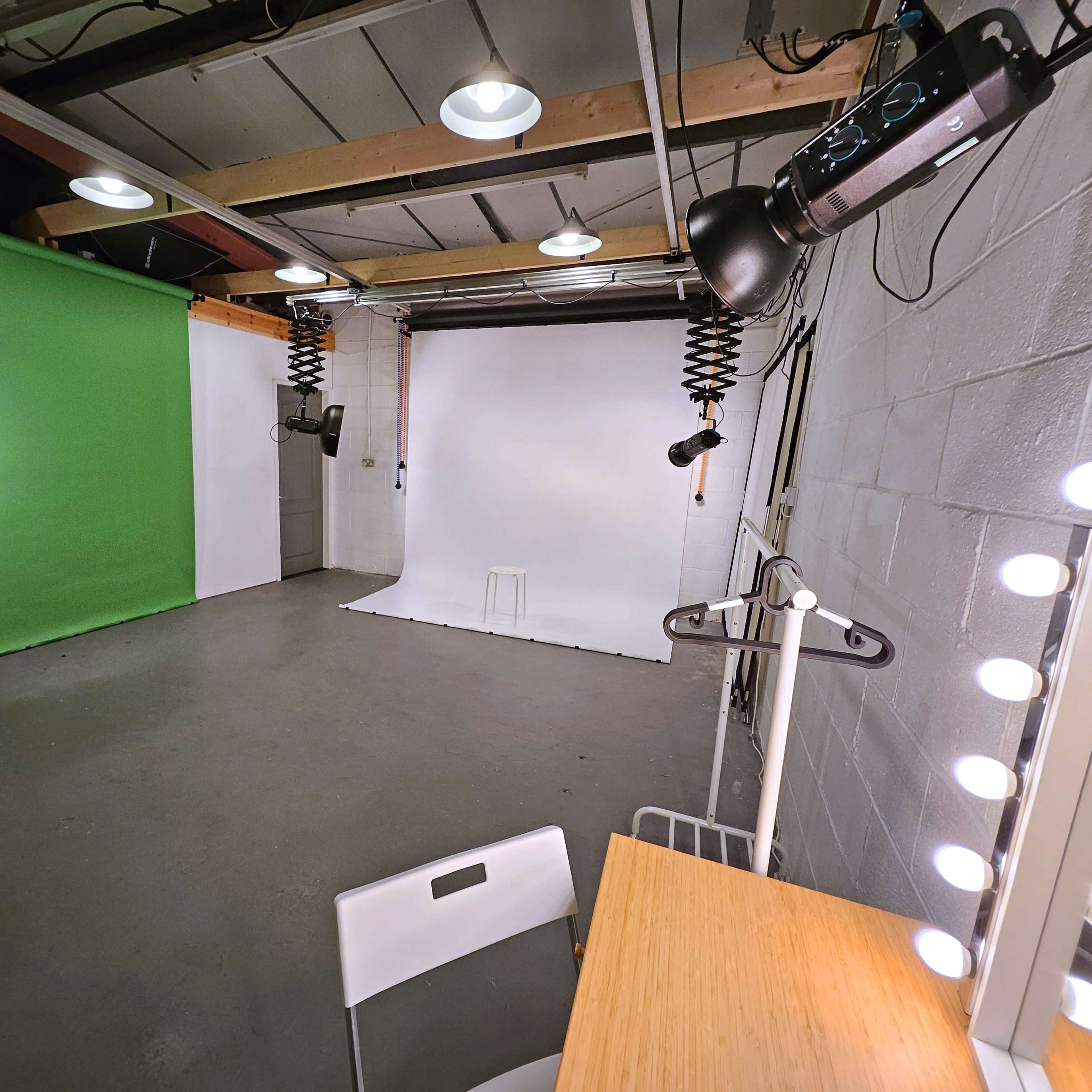 A studio space with a green backdrop on one side, a white backdrop on the other, and a vanity mirror with lights beside a small table and chair.