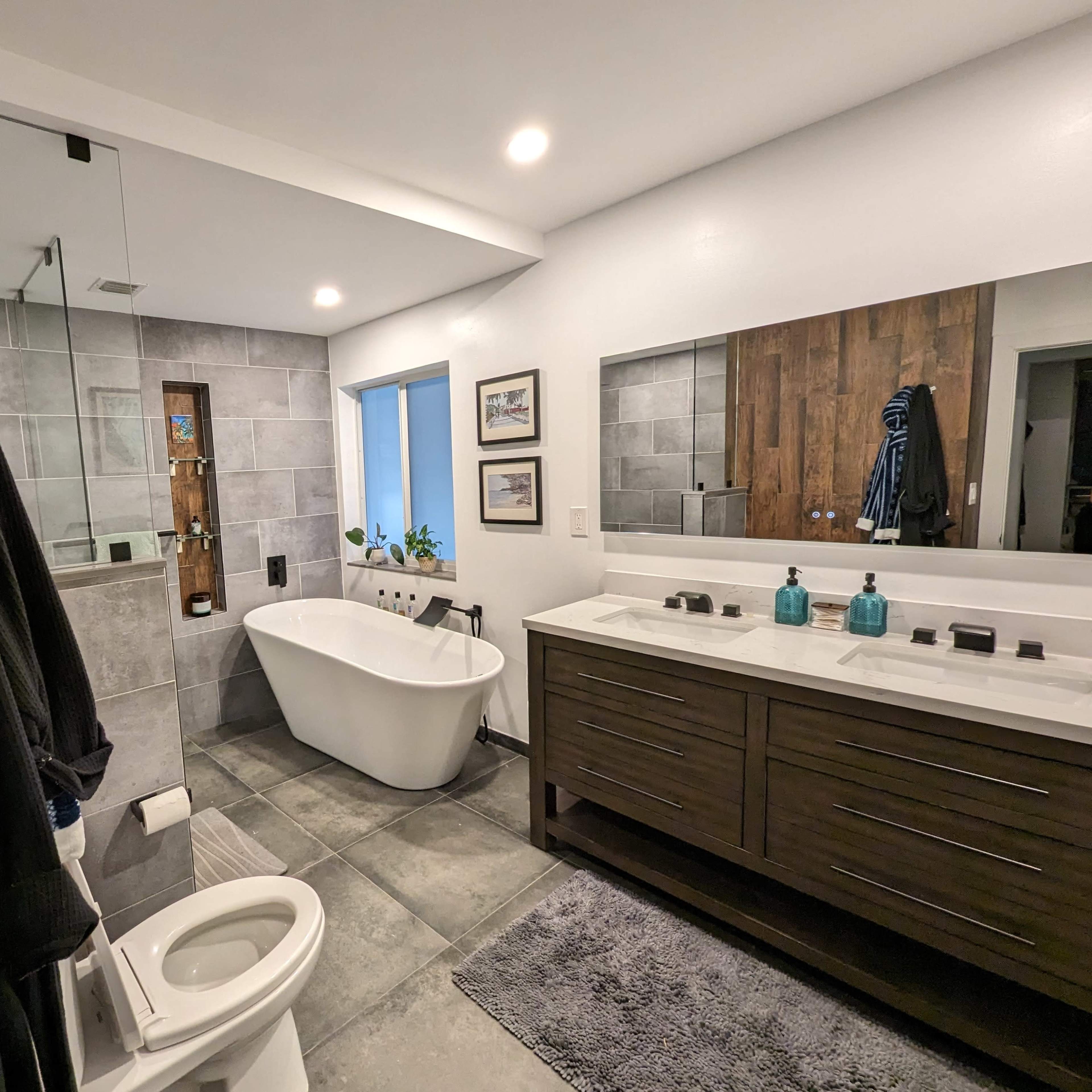 The bathroom features a freestanding tub, double vanity with dark wood cabinetry, a large mirror, and a glass shower enclosure, with gray tiles on the floor and walls.
