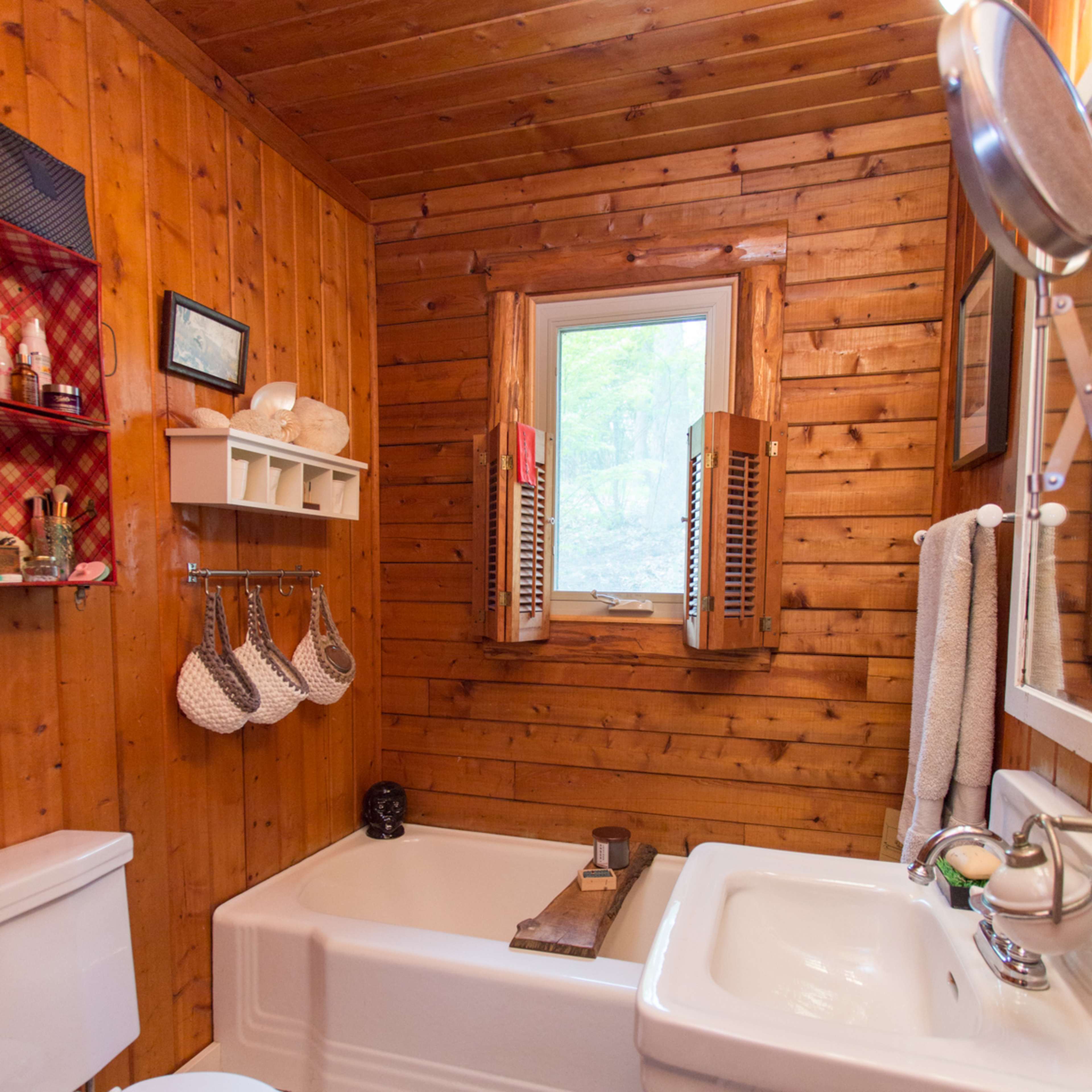 A small, rustic bathroom with wooden walls, a bathtub, a sink, a window, and organized wall storage.