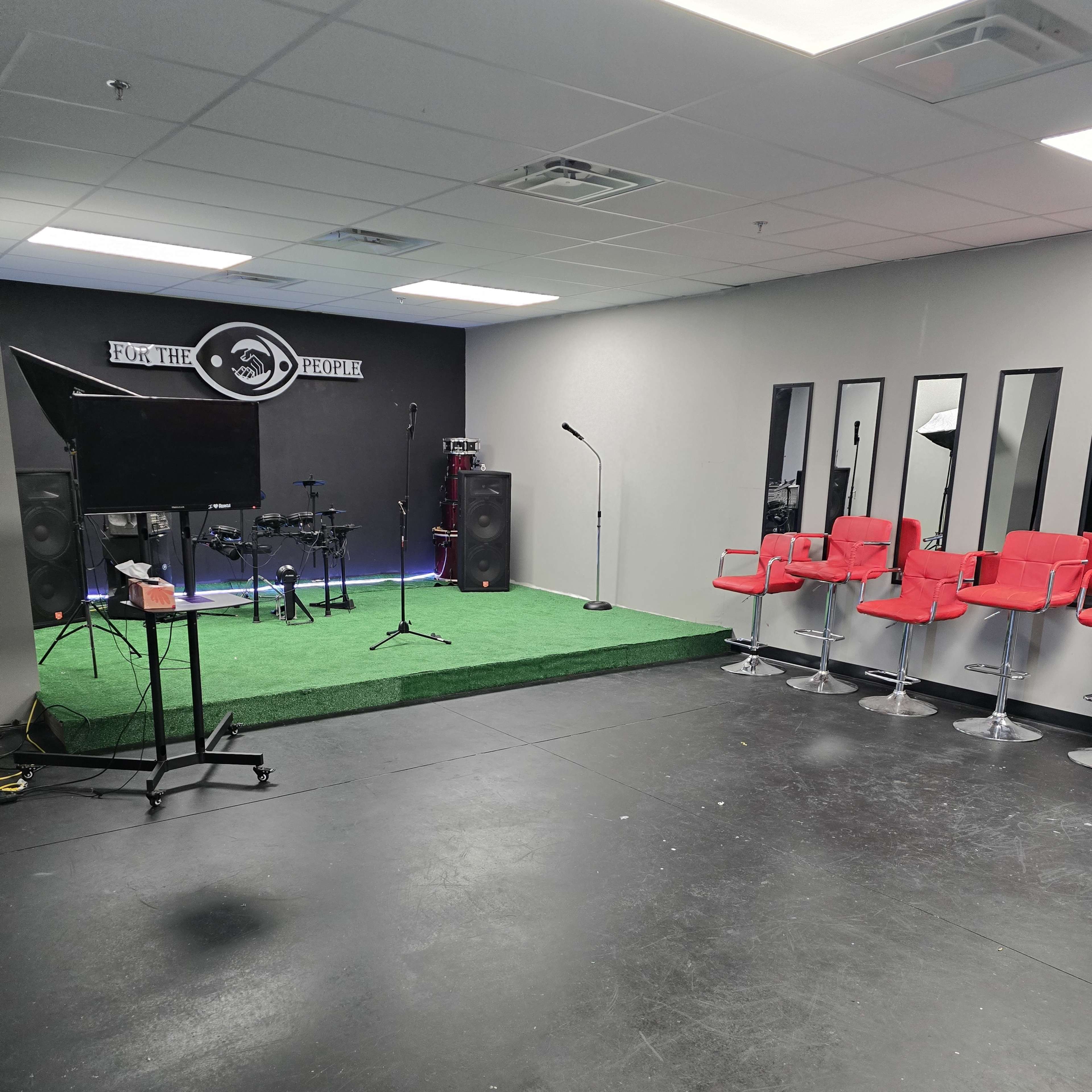 A music studio with a small stage, drum set, and red stools arranged near large mirrors.