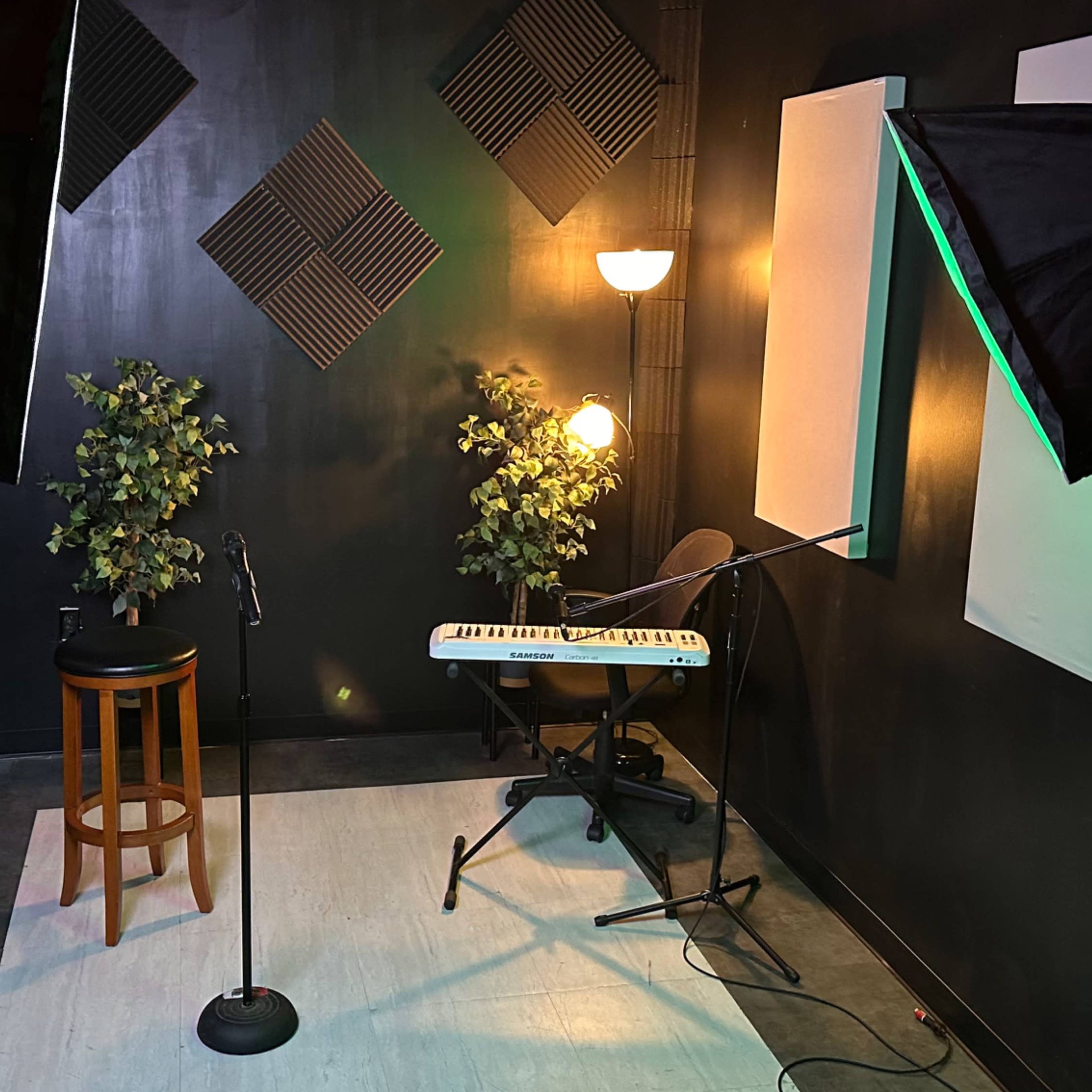 A small recording studio features microphone stands, a keyboard, a stool, and soft lighting against dark walls.