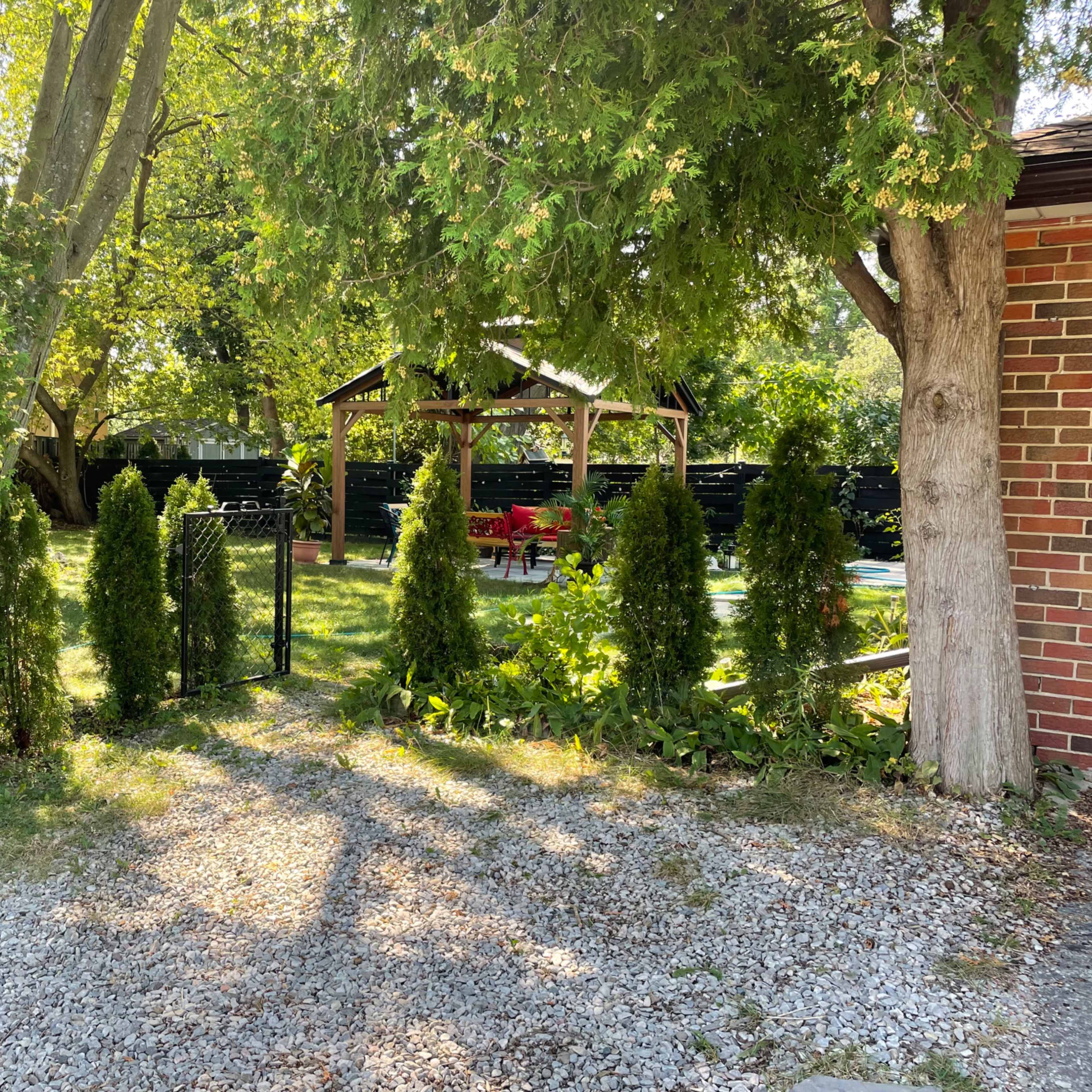 A gravel pathway leads to a fenced backyard with a gazebo and seating area surrounded by greenery.