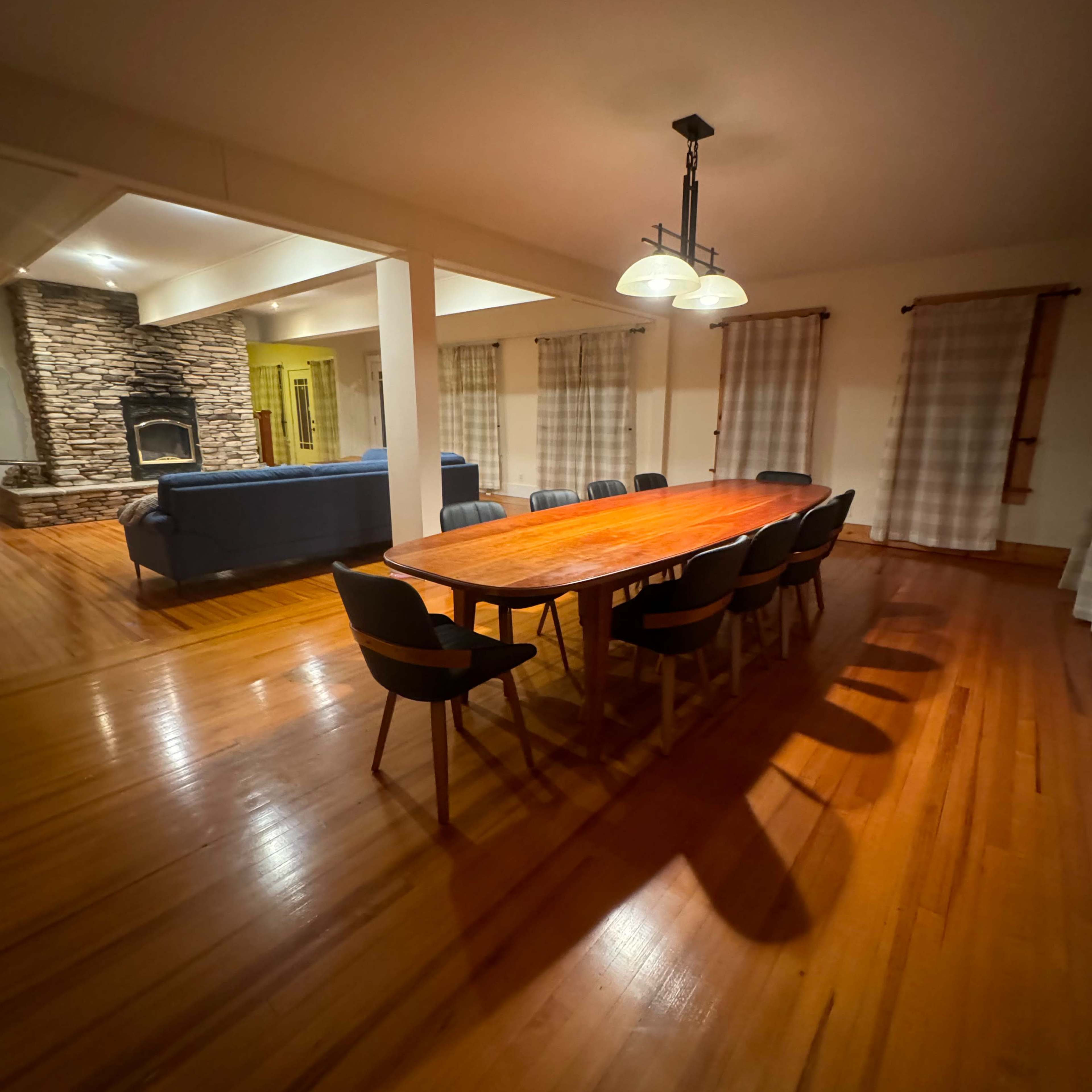 A large wooden dining table with eight chairs is centered in a well-lit room featuring a stone fireplace and a couch in the background.