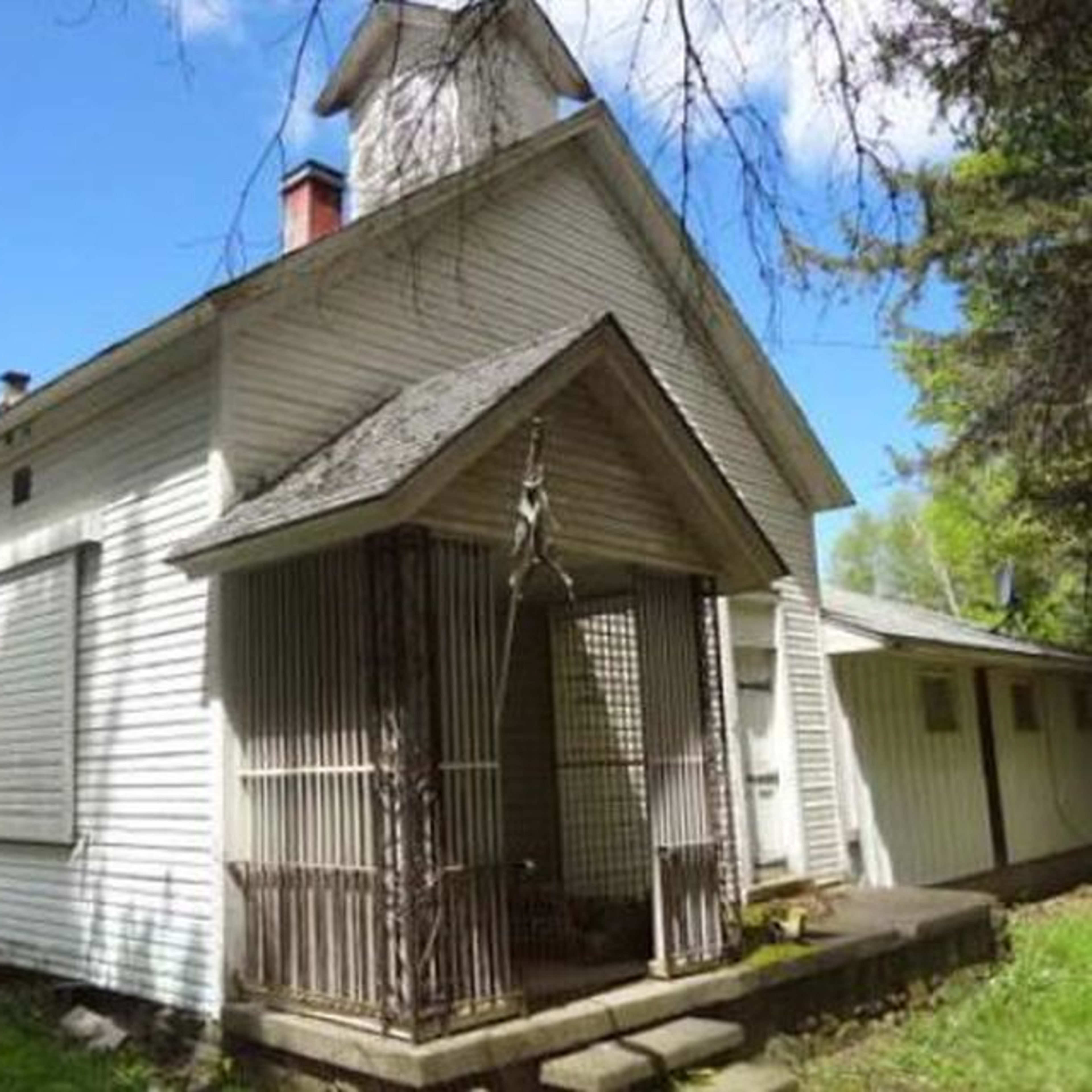 A weathered, single-story white house with a front porch and a bell tower, surrounded by grass and trees.