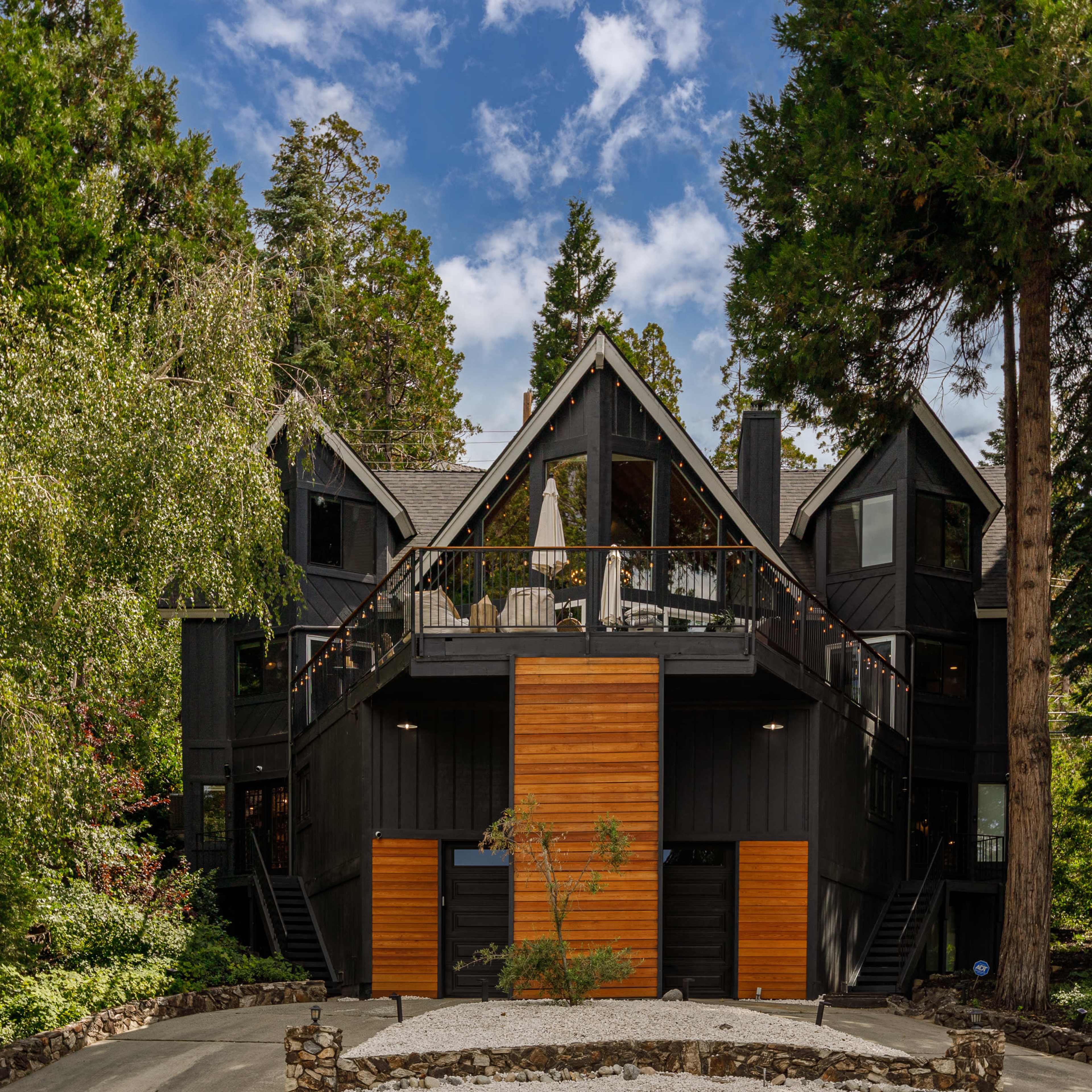 A modern black chalet-style house with wooden accents is surrounded by tall trees and greenery.