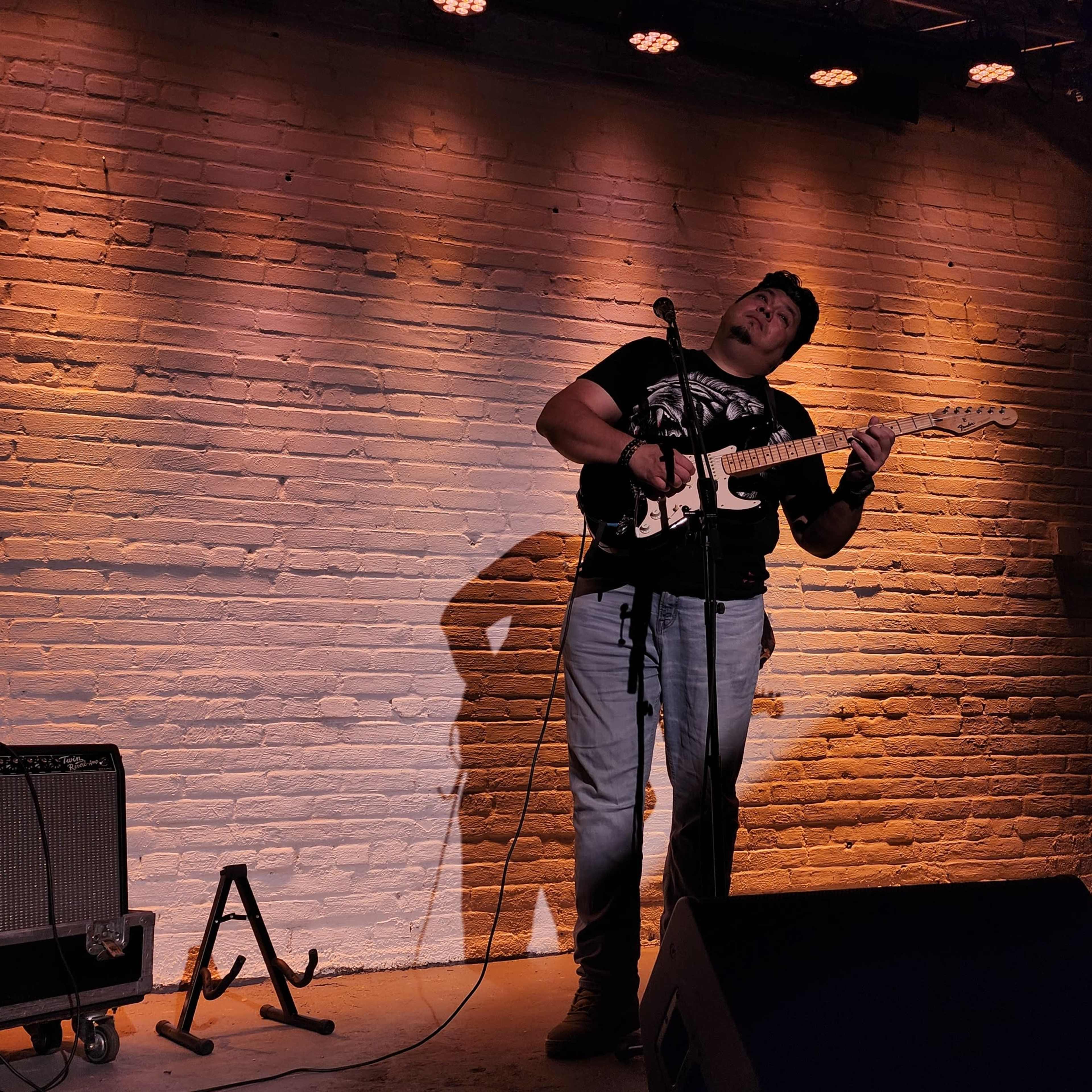 A musician plays an electric guitar on stage in front of a brick wall, with spotlights illuminating the scene.