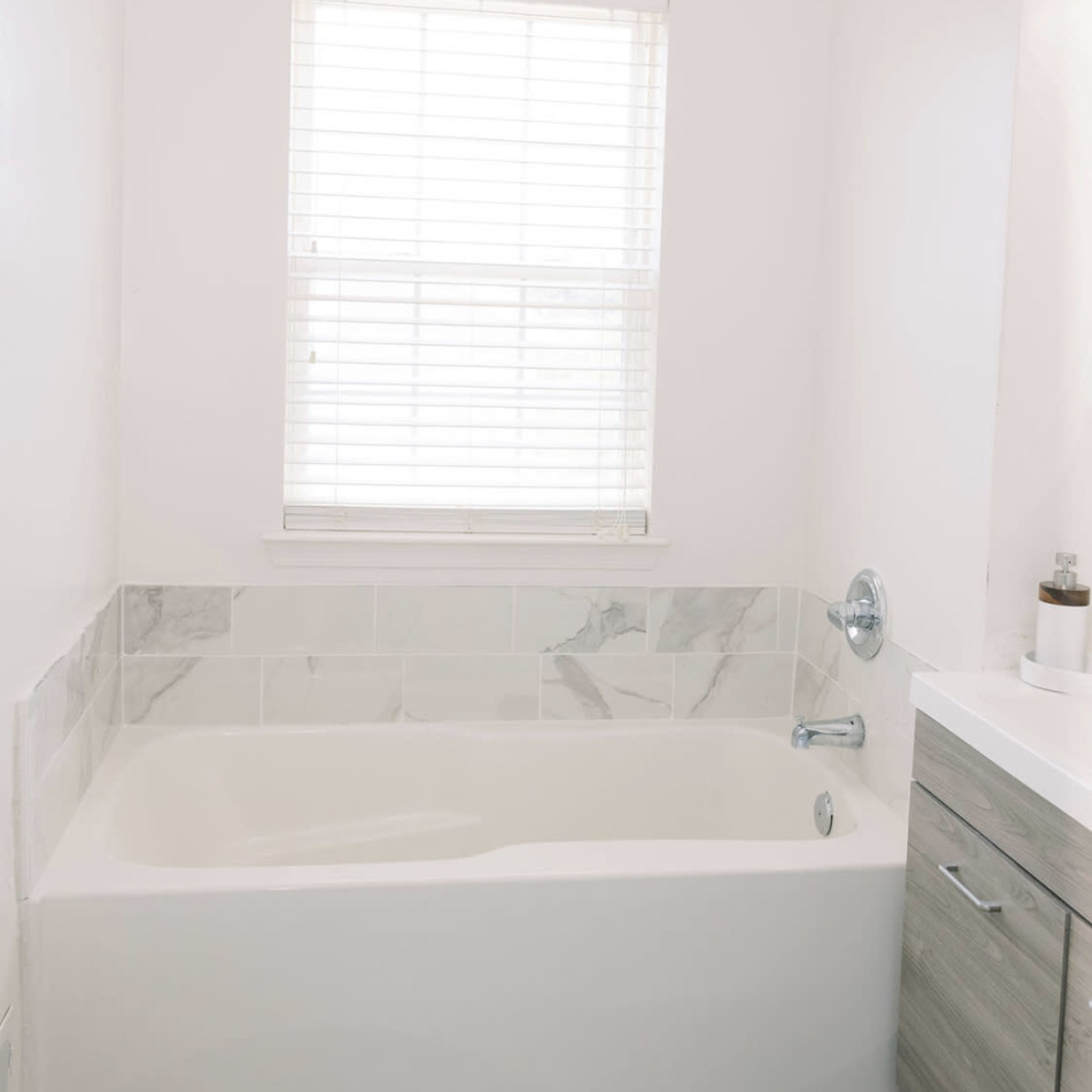 A bathroom featuring a white bathtub with a marble accent wall, a window with blinds, and a gray cabinet.