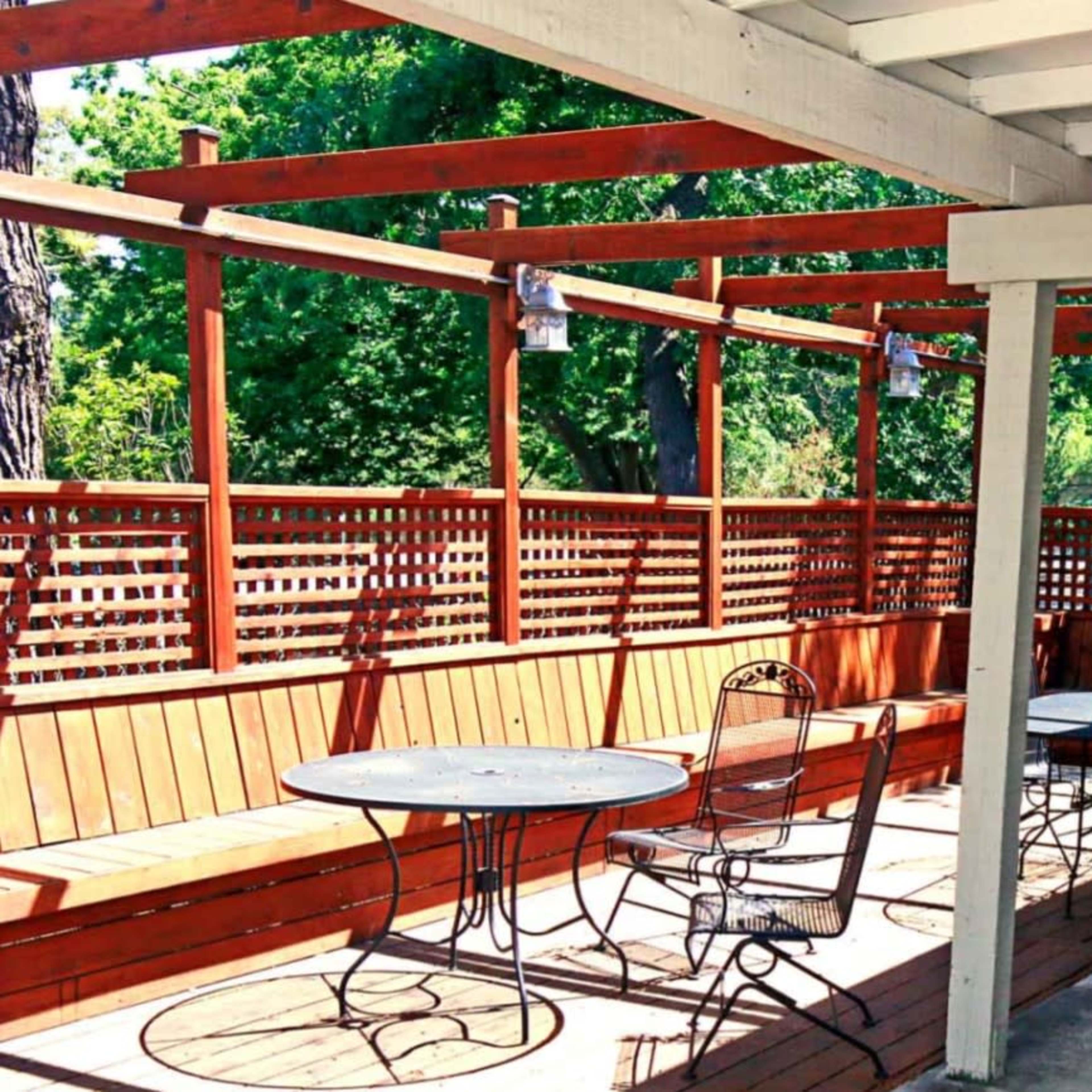 A patio area with wooden benches and round tables, surrounded by wooden lattice and lush greenery.