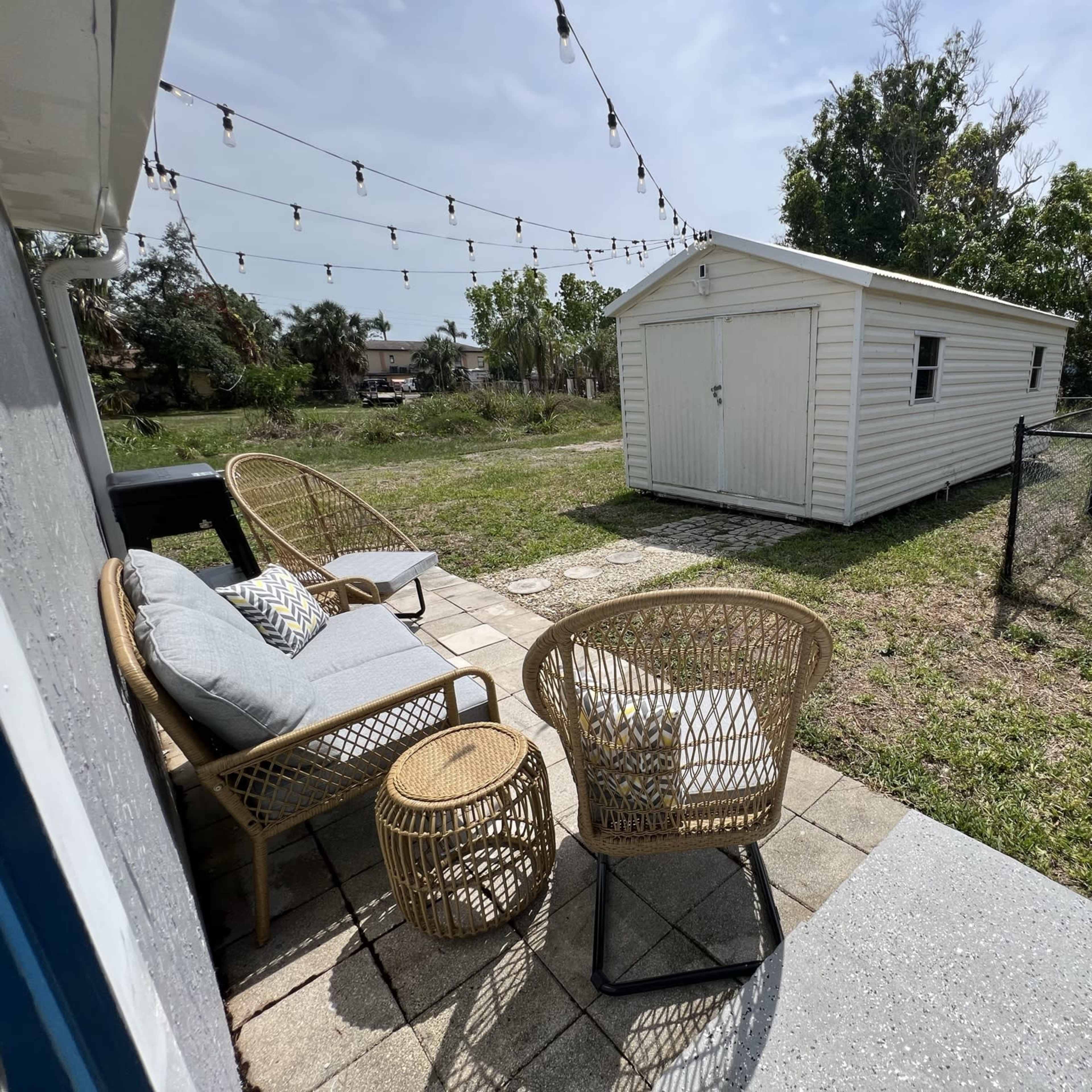 A patio area features two wicker chairs and a small table, with a white shed in the background and string lights overhead.