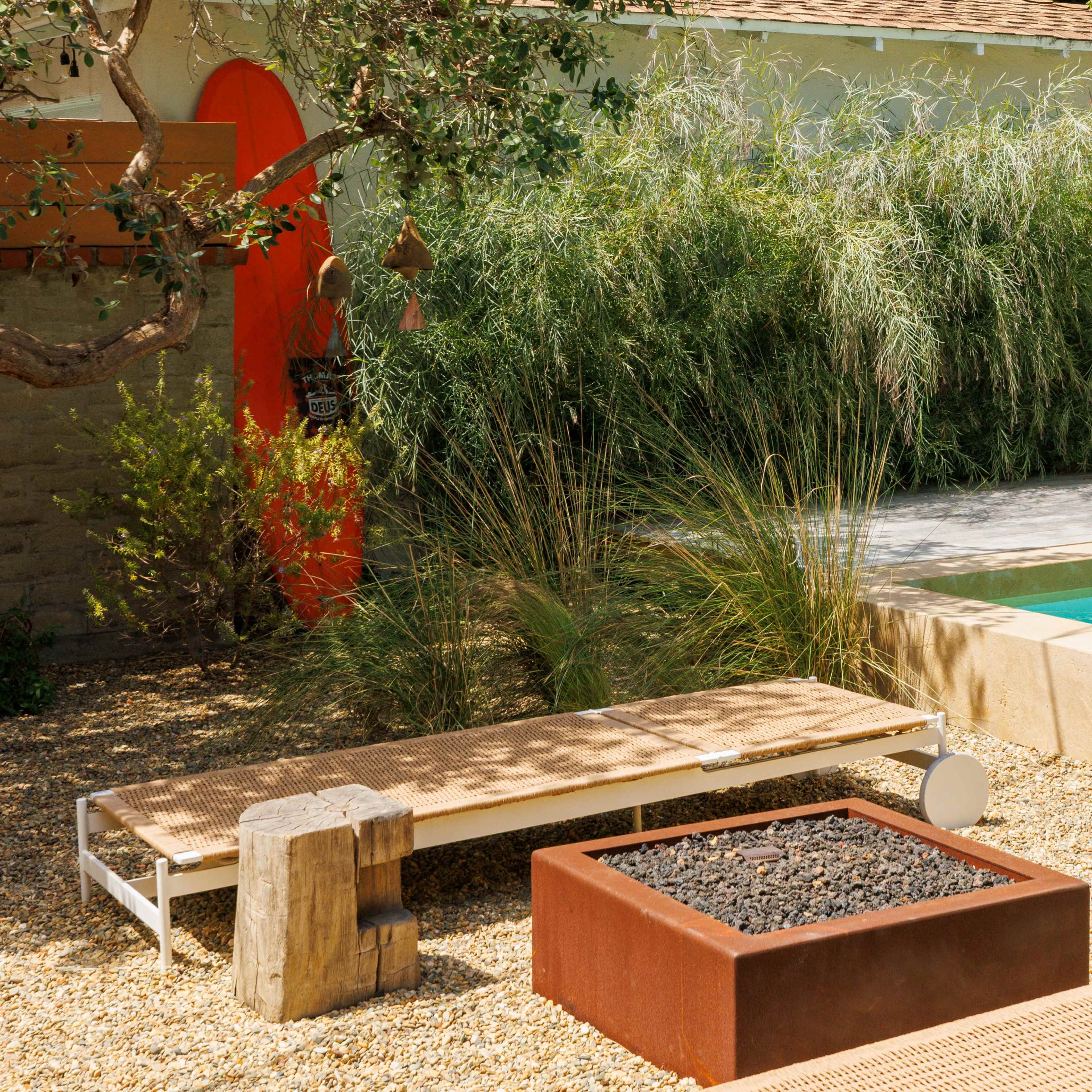 The scene features a landscaped backyard with a swimming pool, a fire pit surrounded by gravel, and lounge chairs near tall grasses and an orange door in the background.