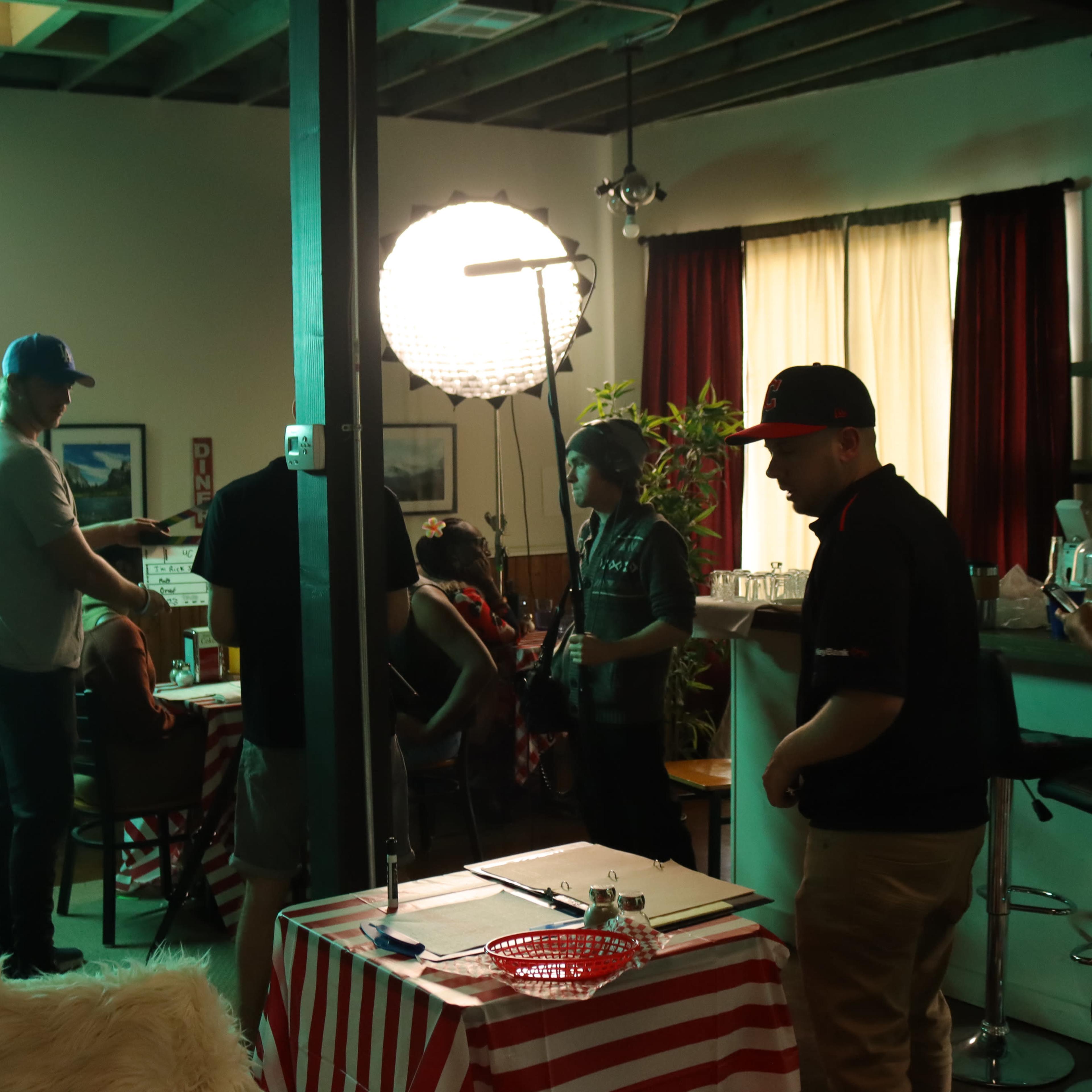 A group of people is gathered in a dimly lit room with bar stools and a round light source, while some are seated at a table and others stand nearby.