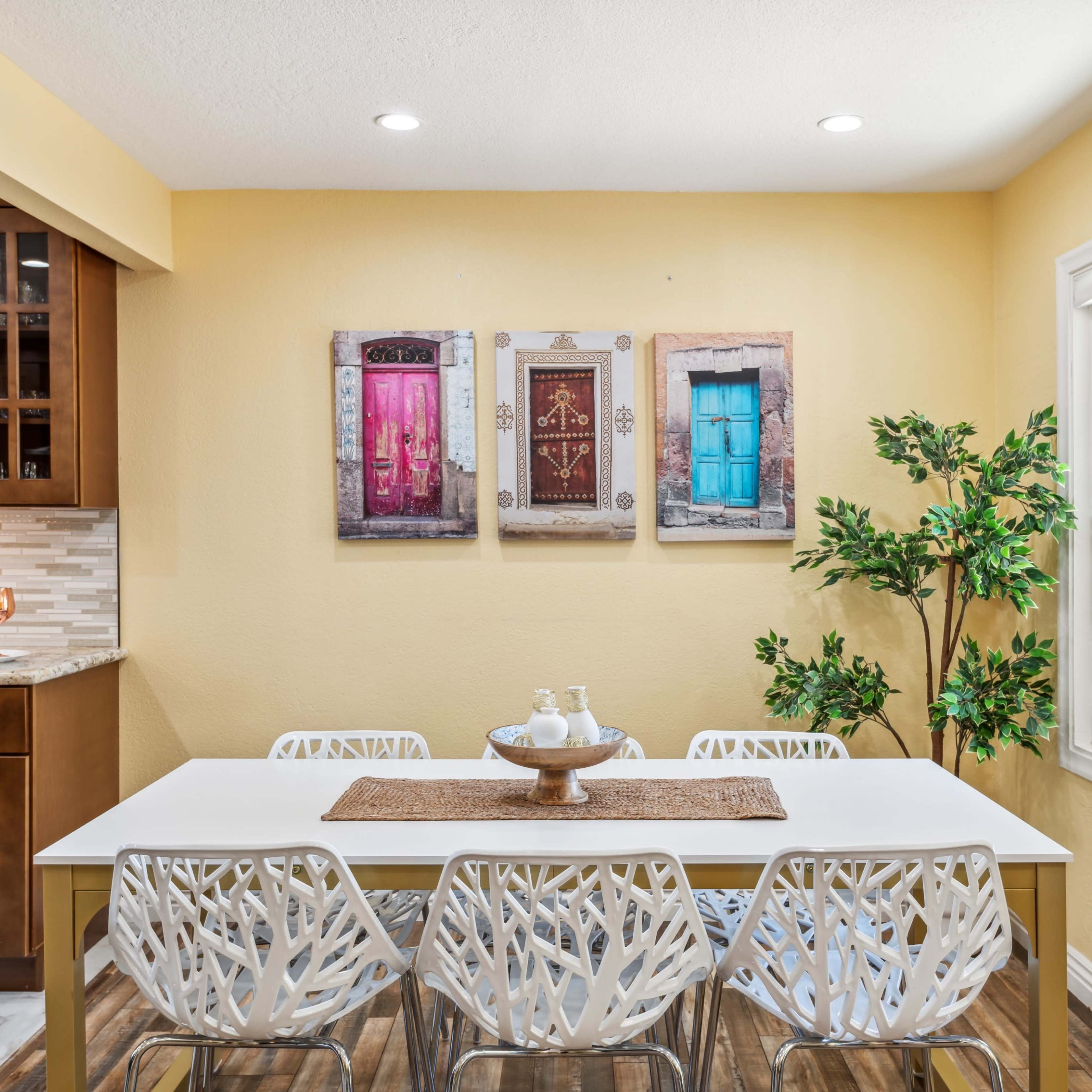 A dining area features a white table surrounded by modern chairs, with framed colorful door artwork on the wall and a small plant in the corner.