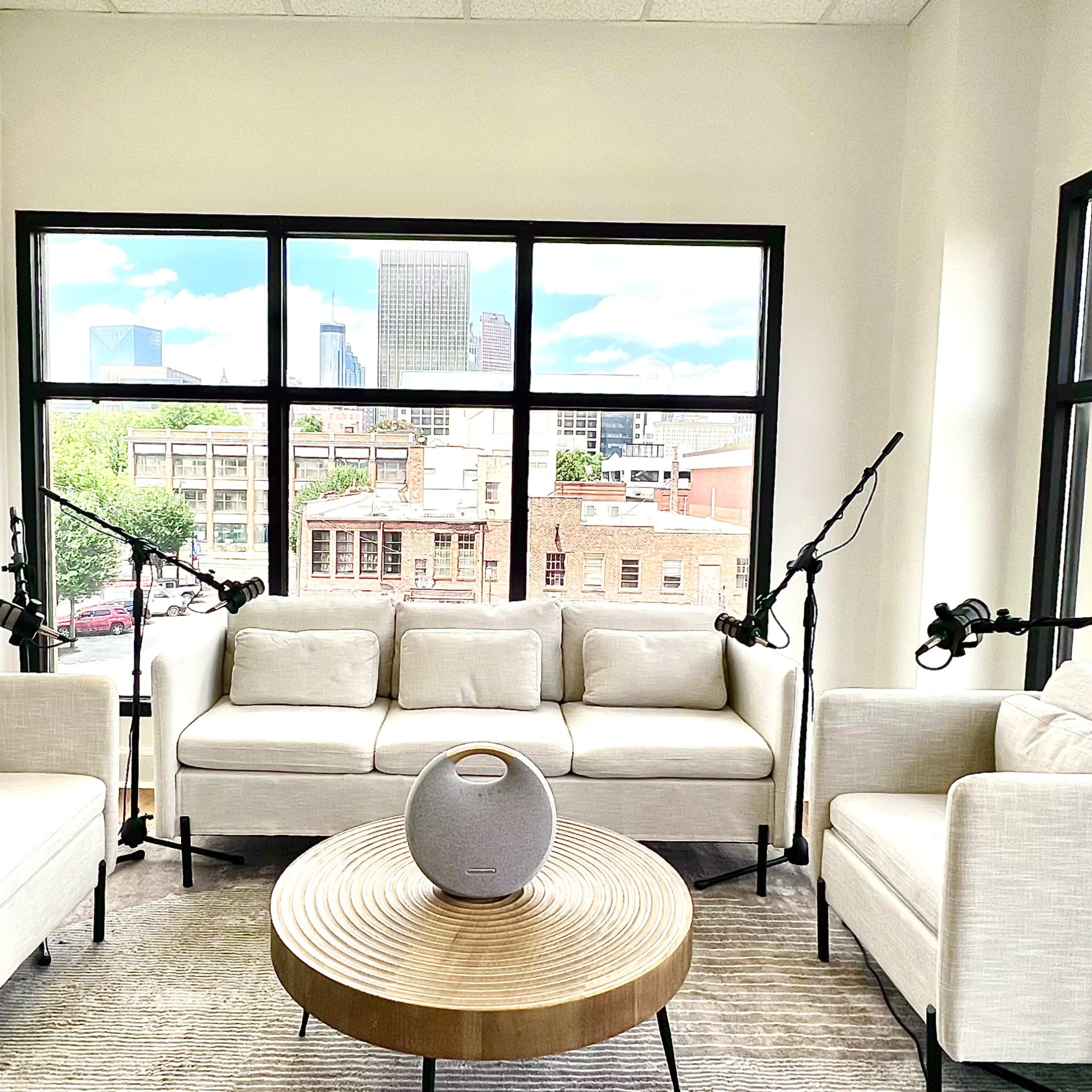 A bright interior features two white sofas and a round coffee table near large windows overlooking a cityscape.