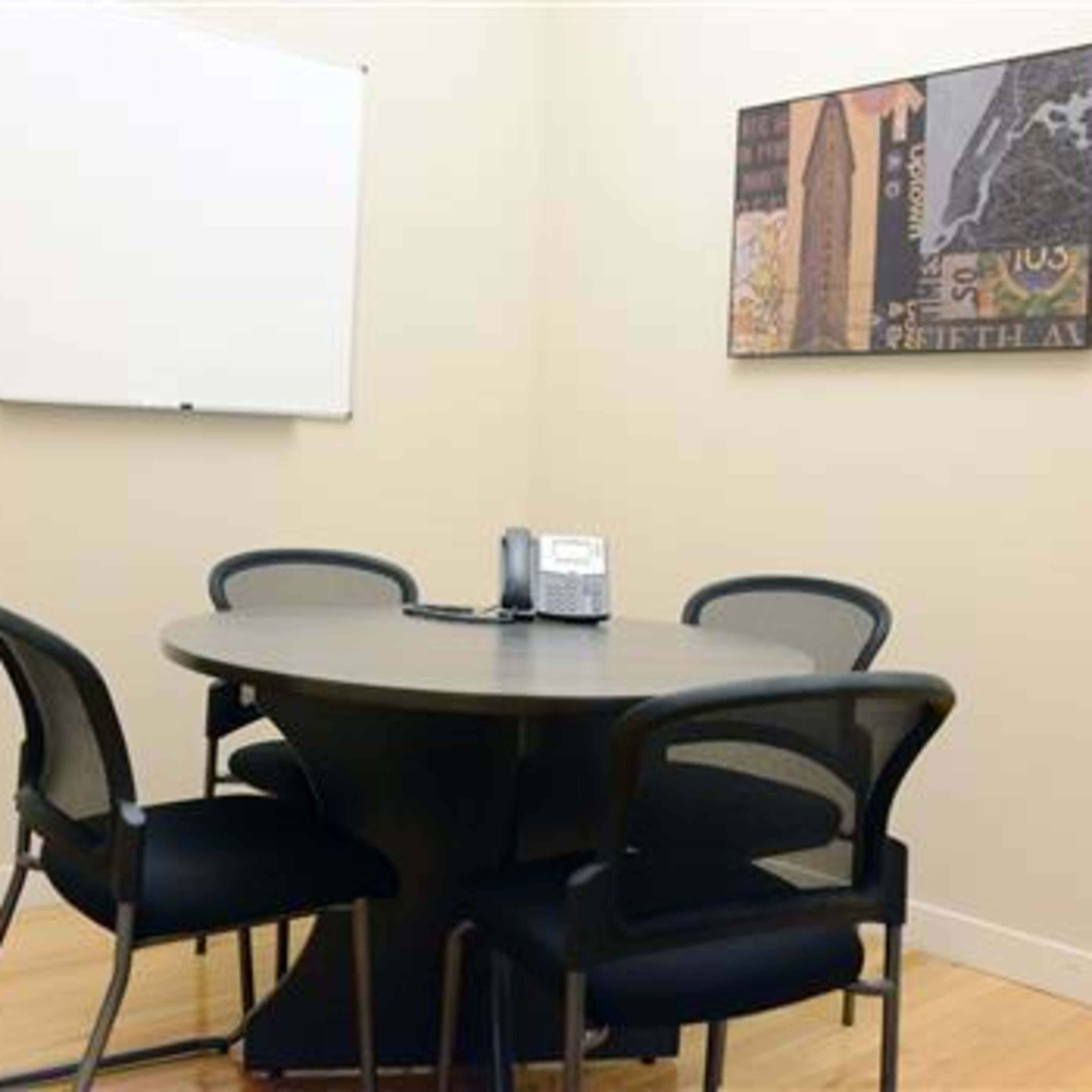A small conference room features a round table with five black chairs, a telephone, and a whiteboard on the wall.