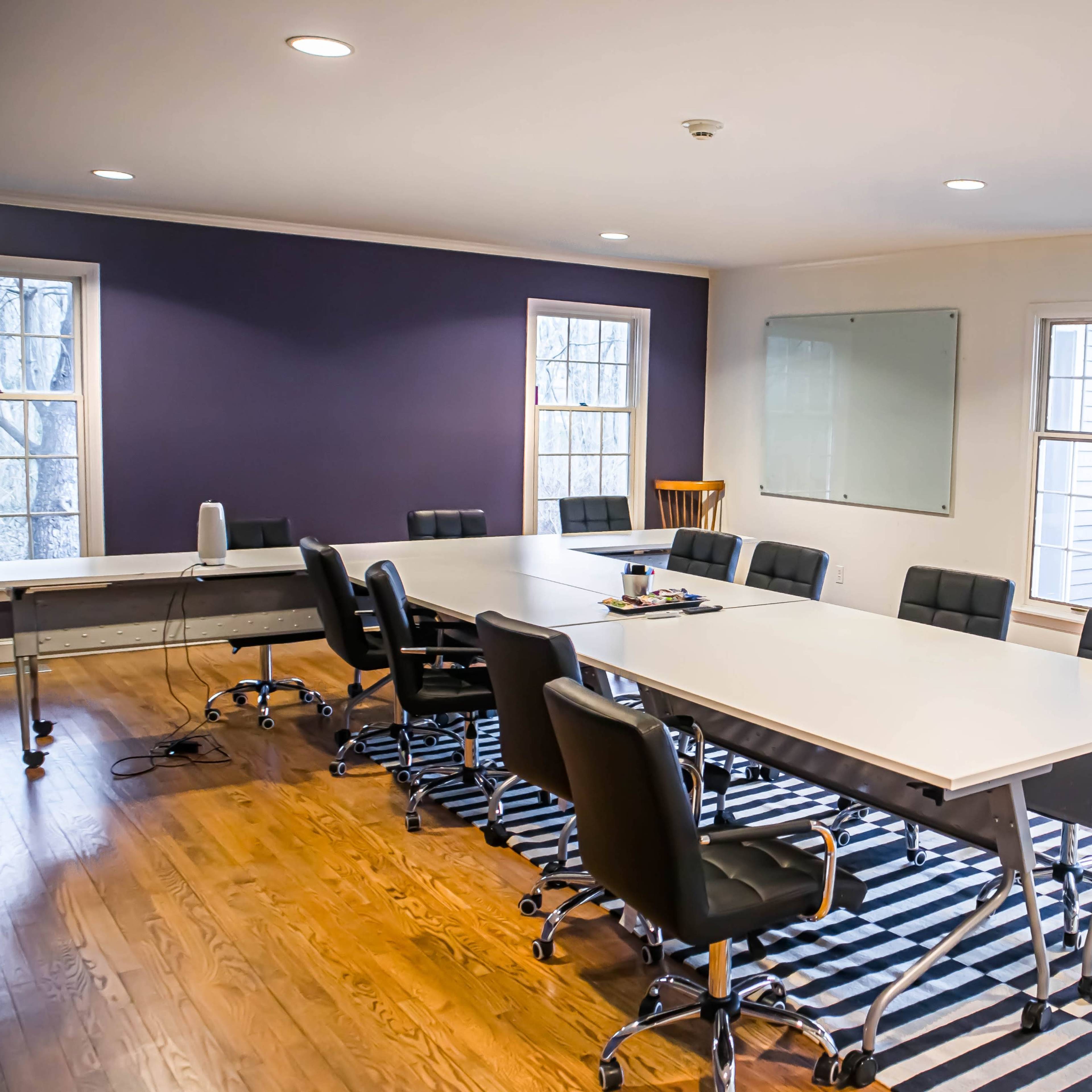 A spacious conference room features a long table surrounded by black rolling chairs, with large windows allowing natural light to illuminate the space.