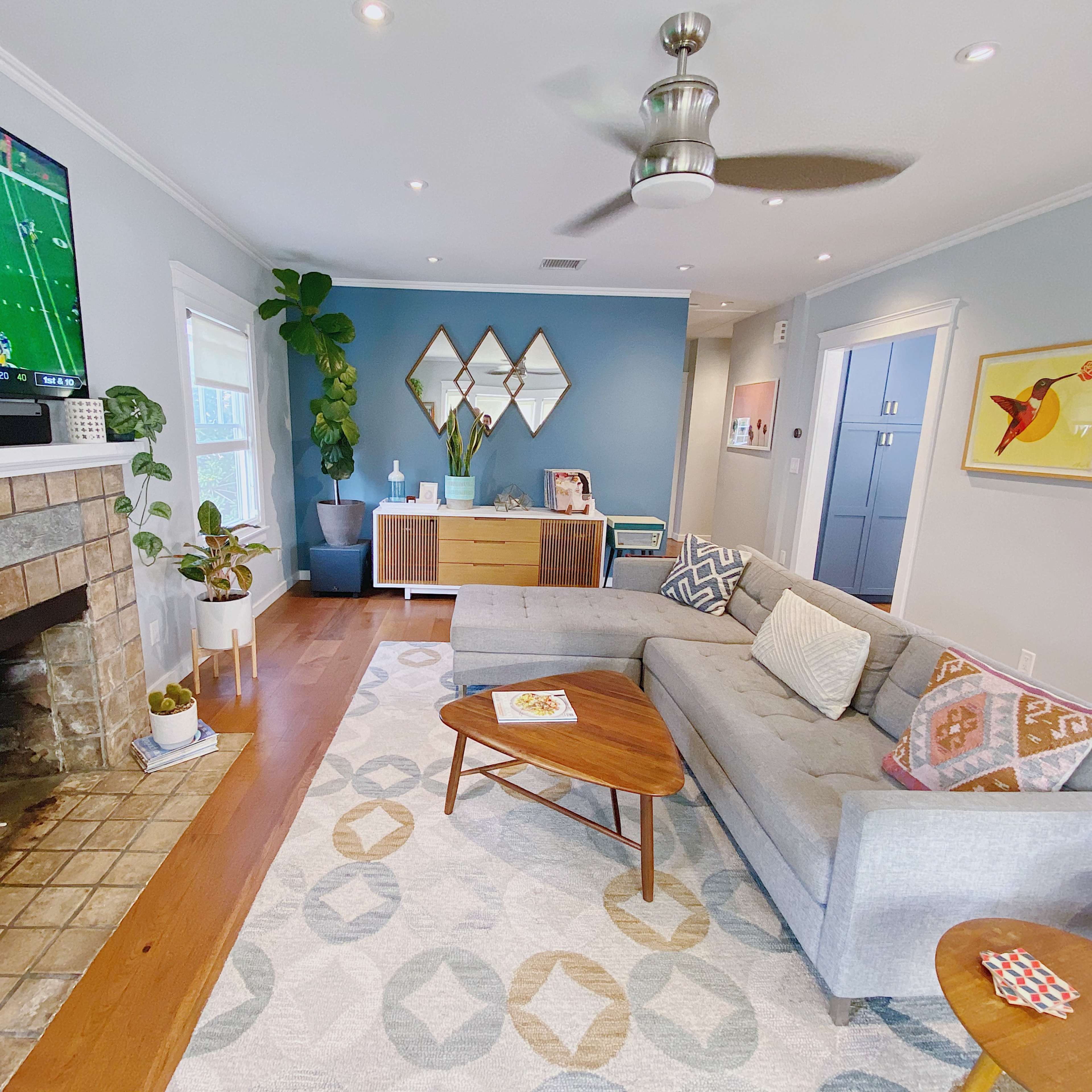 A modern living room features a gray sectional sofa, a stone fireplace, a television displaying a football game, and decorative plants.