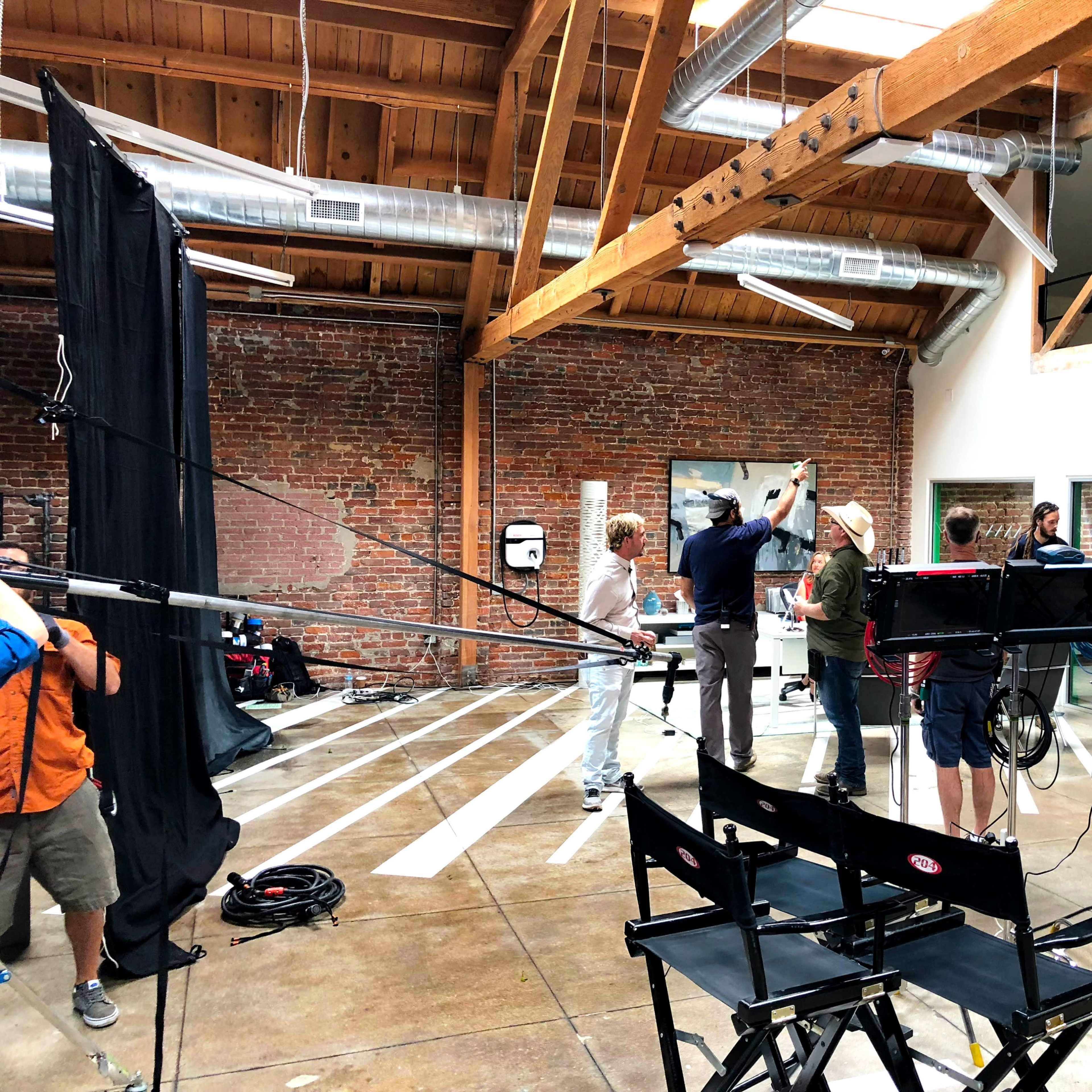 A film crew is setting up equipment in a spacious studio with exposed brick walls and wooden beams.