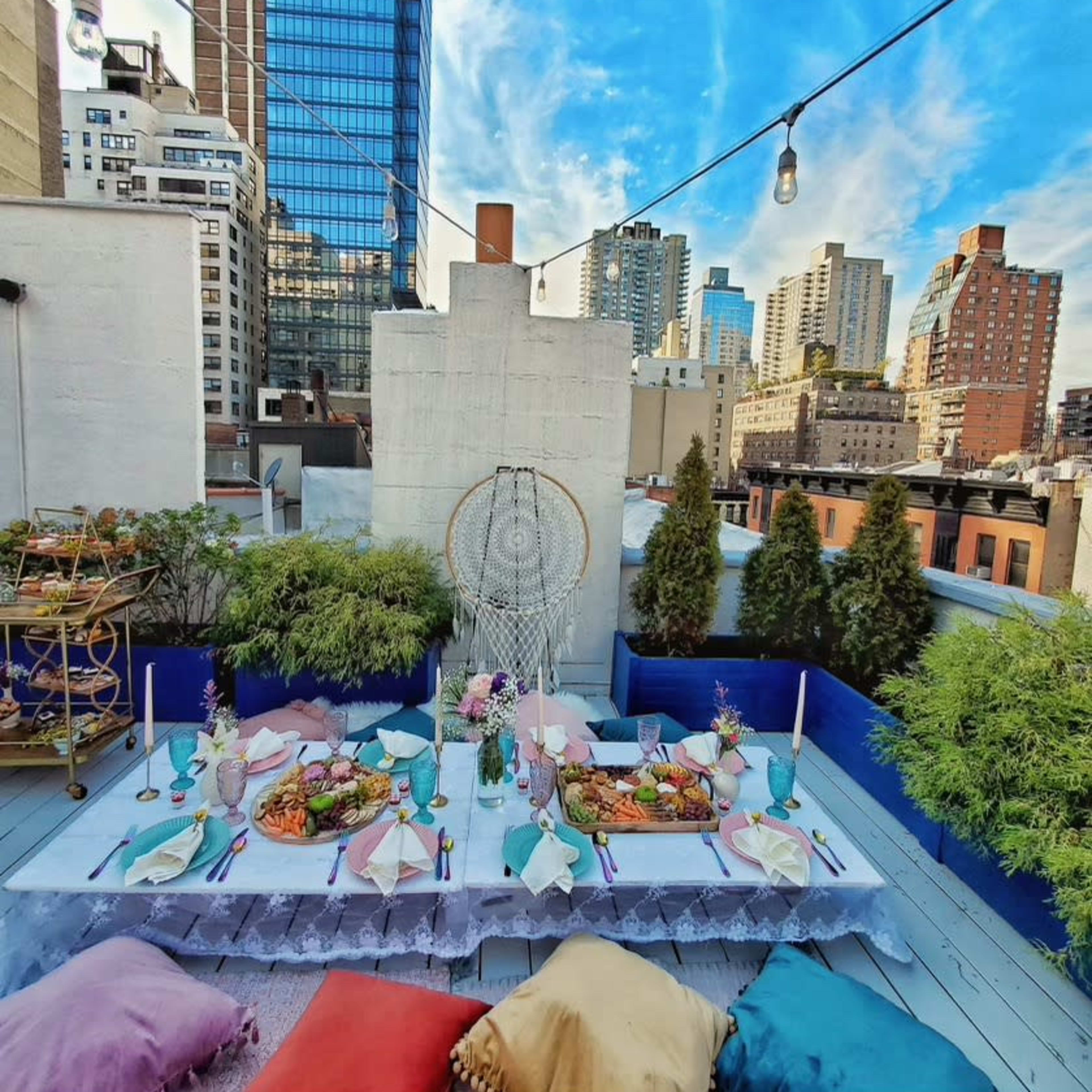 A decorated rooftop terrace features a long dining table surrounded by colorful cushions and city skyscrapers in the background.