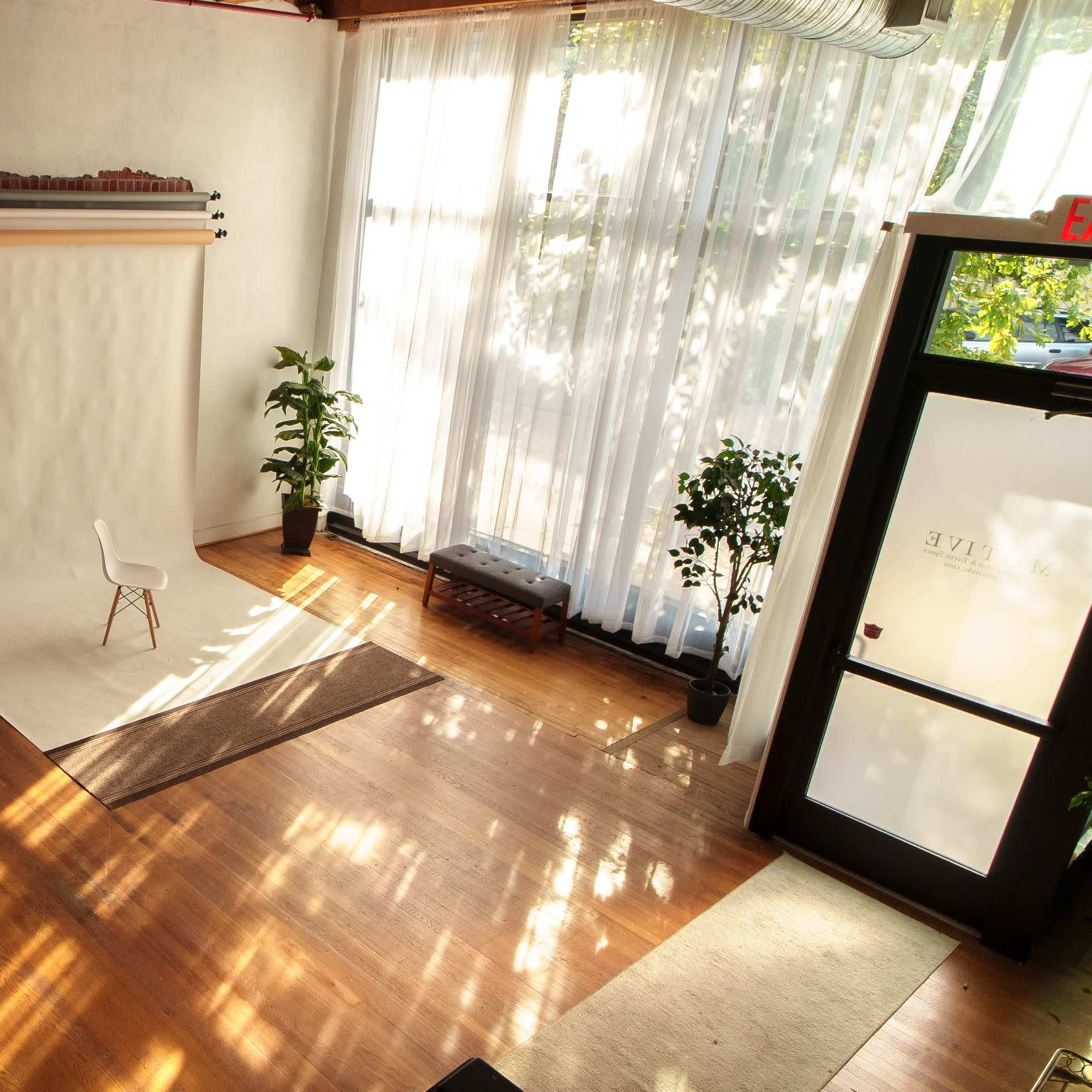 A bright studio space with a white backdrop, a single chair in front, wooden flooring, large windows letting in natural light, and potted plants.