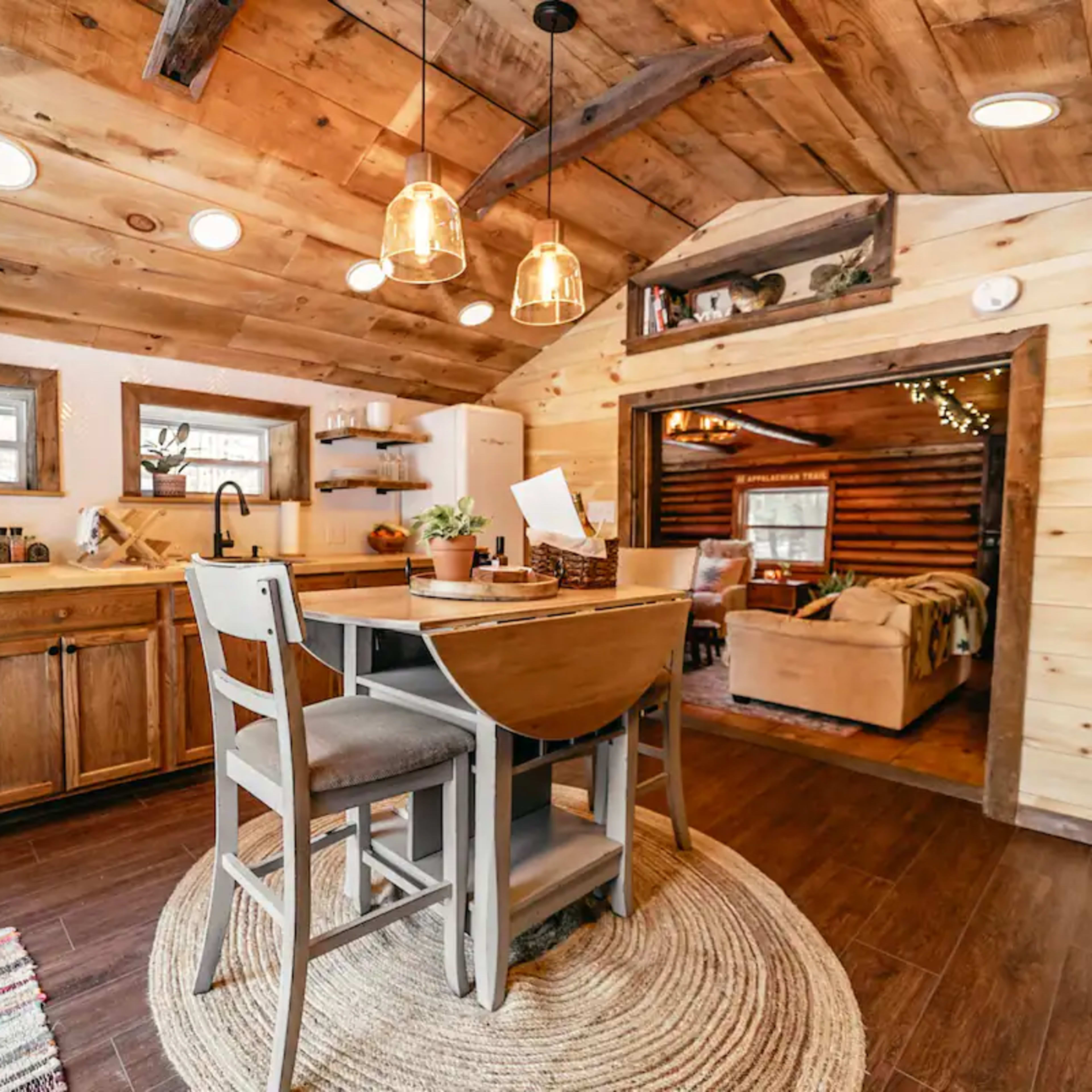 A cozy kitchen with a wooden ceiling, a dining table for two, and a living area visible through an archway.