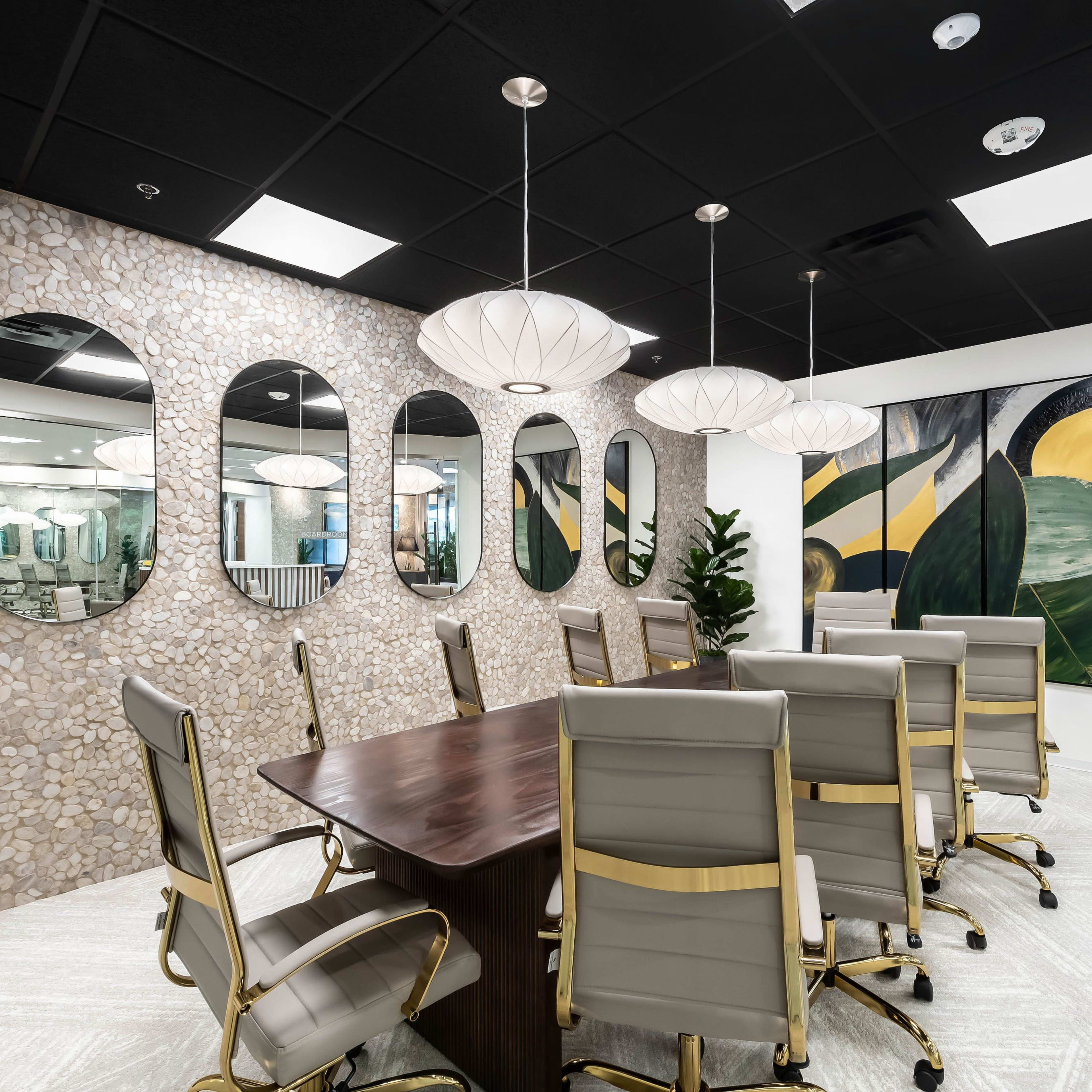 A modern conference room features a long wooden table surrounded by gray leather chairs, with a textured stone wall and several round mirrors, illuminated by pendant lights.