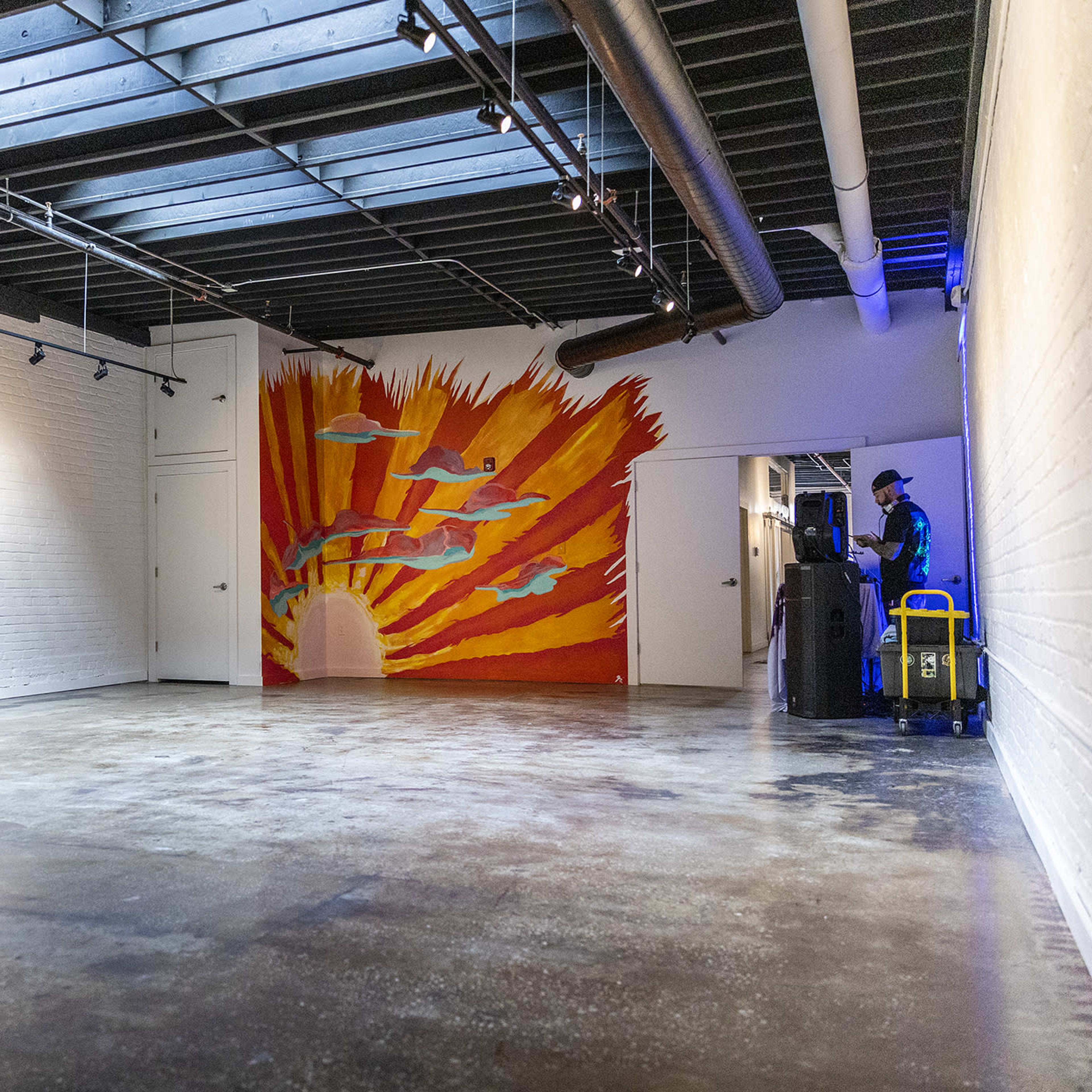 An empty interior space with white brick walls, a painted mural of a sunburst on one wall, and a person standing near a cart in the corner.