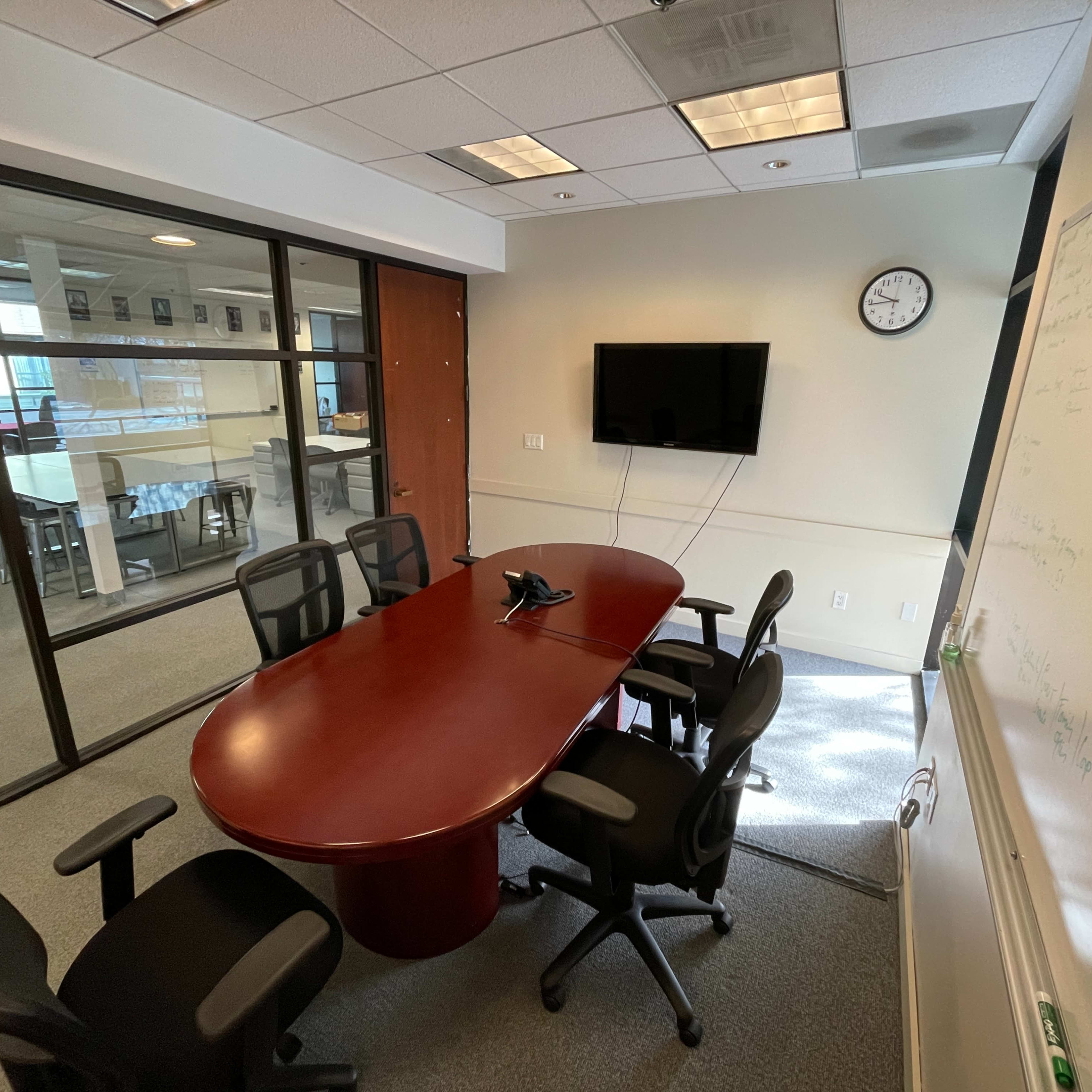A conference room features a large oval table surrounded by black chairs, a wall-mounted television, and a whiteboard.
