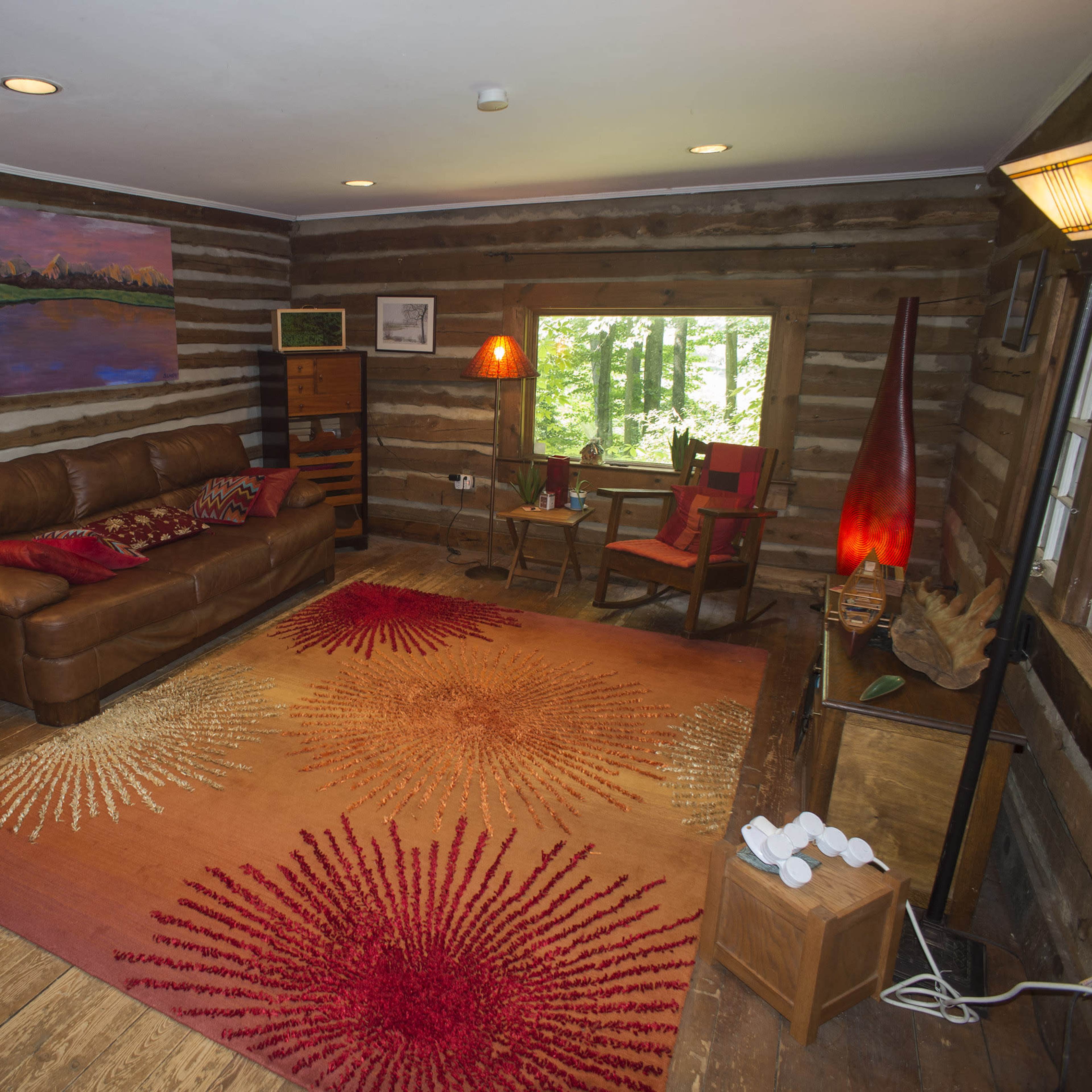 A rustic cabin interior with a leather sofa, wooden furniture, and a bright area rug, surrounded by log walls and a window overlooking trees.
