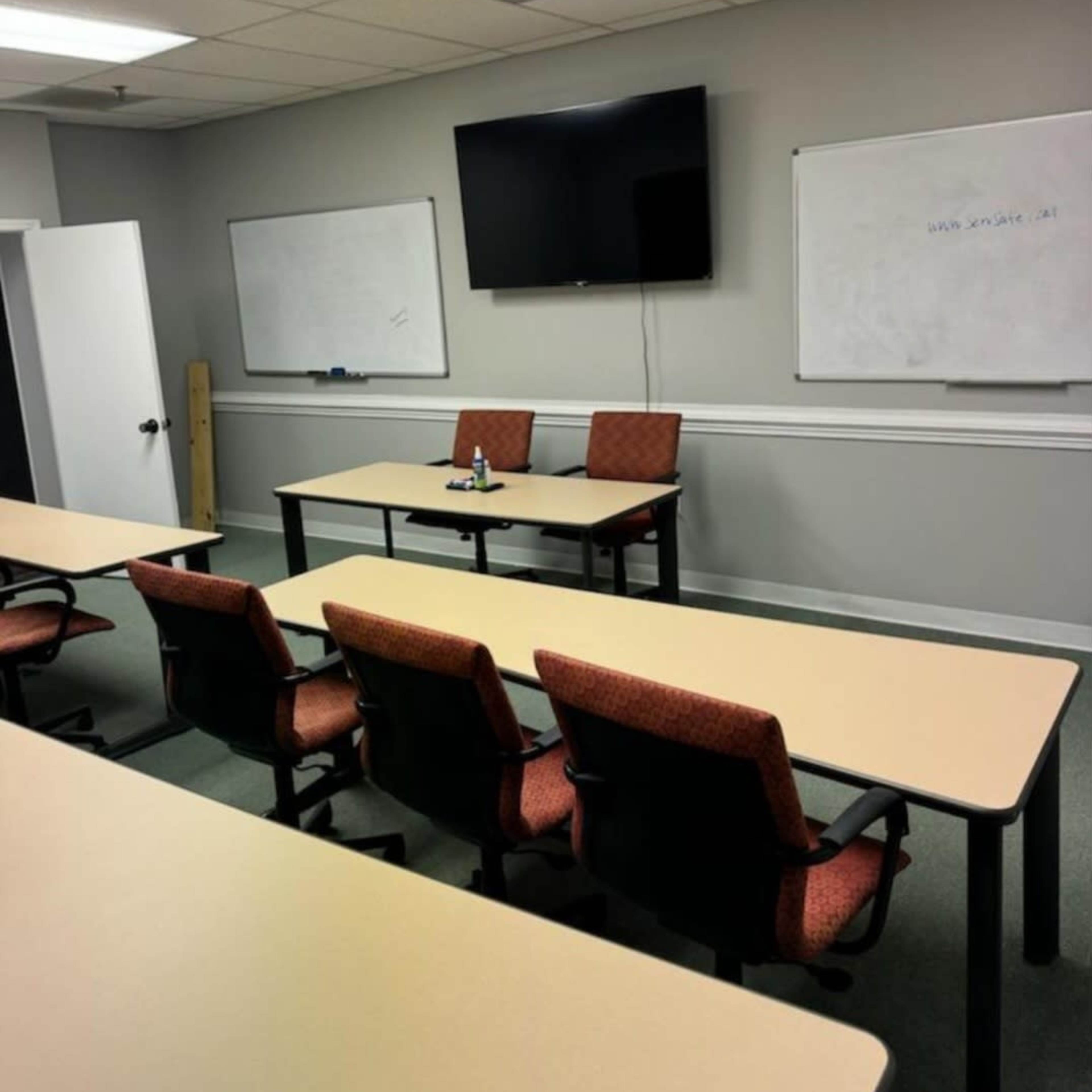 A classroom with rows of chairs and tables facing a television mounted on the wall, and two empty chairs at the front.