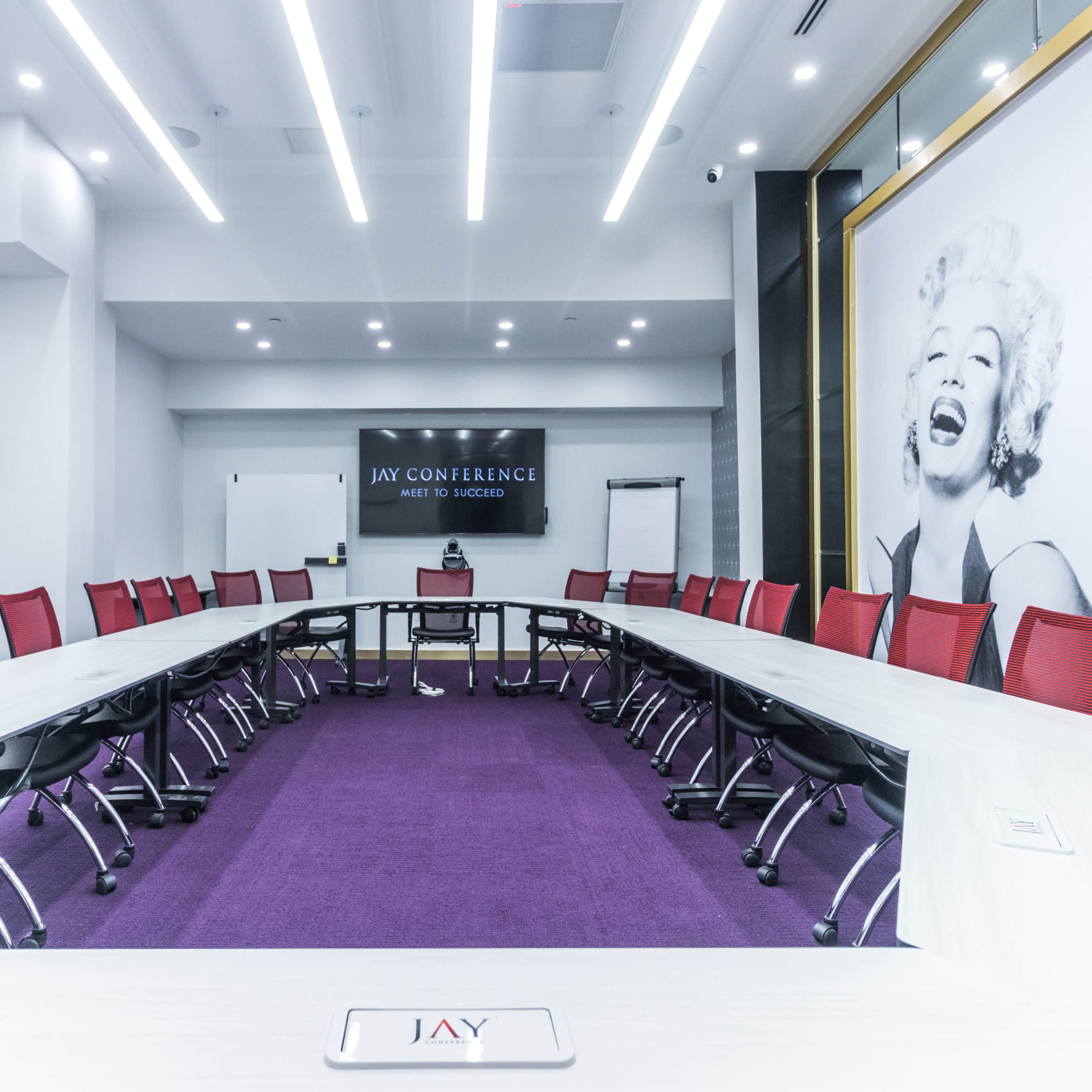 A modern conference room features a large rectangular table surrounded by red chairs, with a purple carpet and a prominent portrait of Marilyn Monroe on the wall.