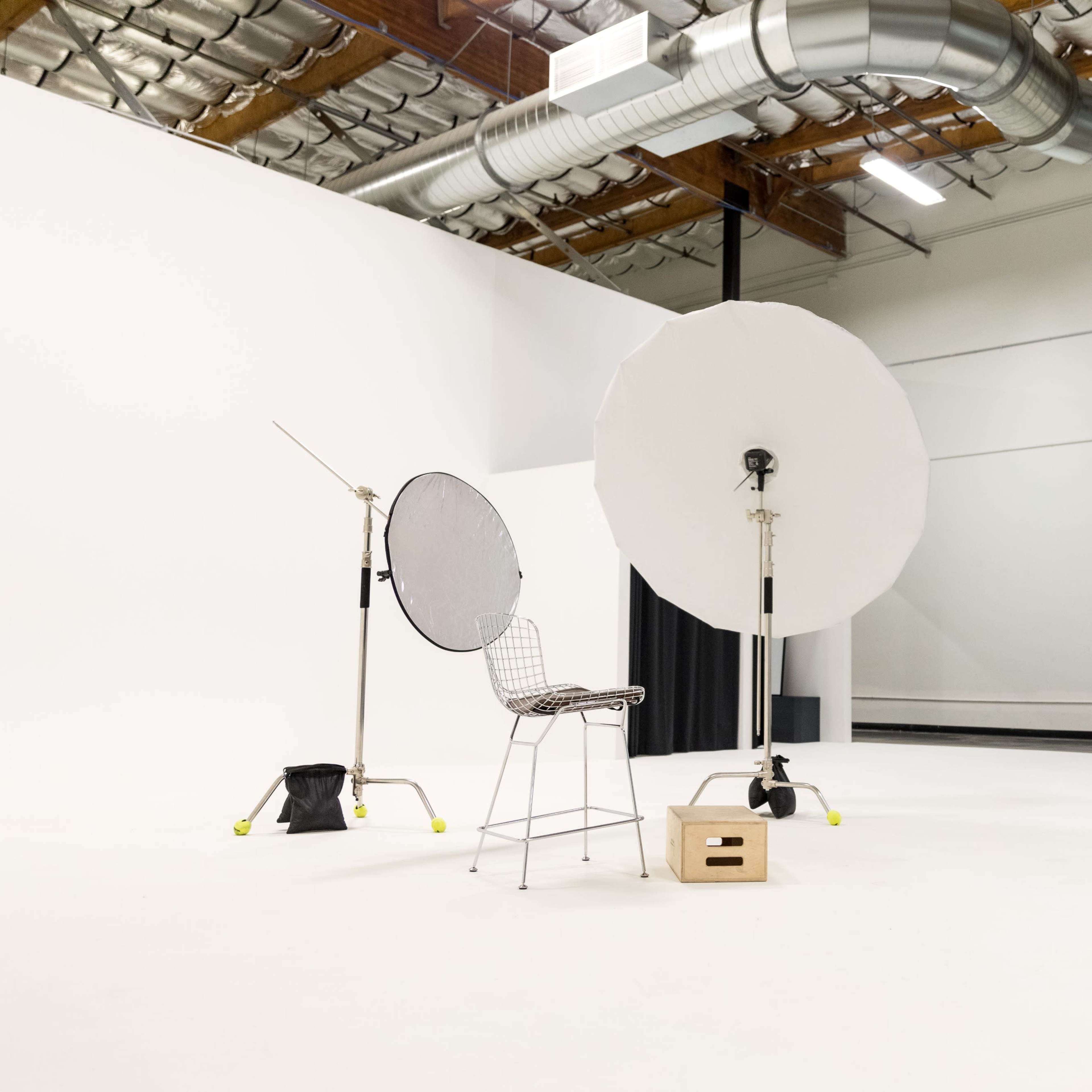 A photography studio features a wire mesh chair surrounded by large light modifiers and a wooden box on a blank white floor.