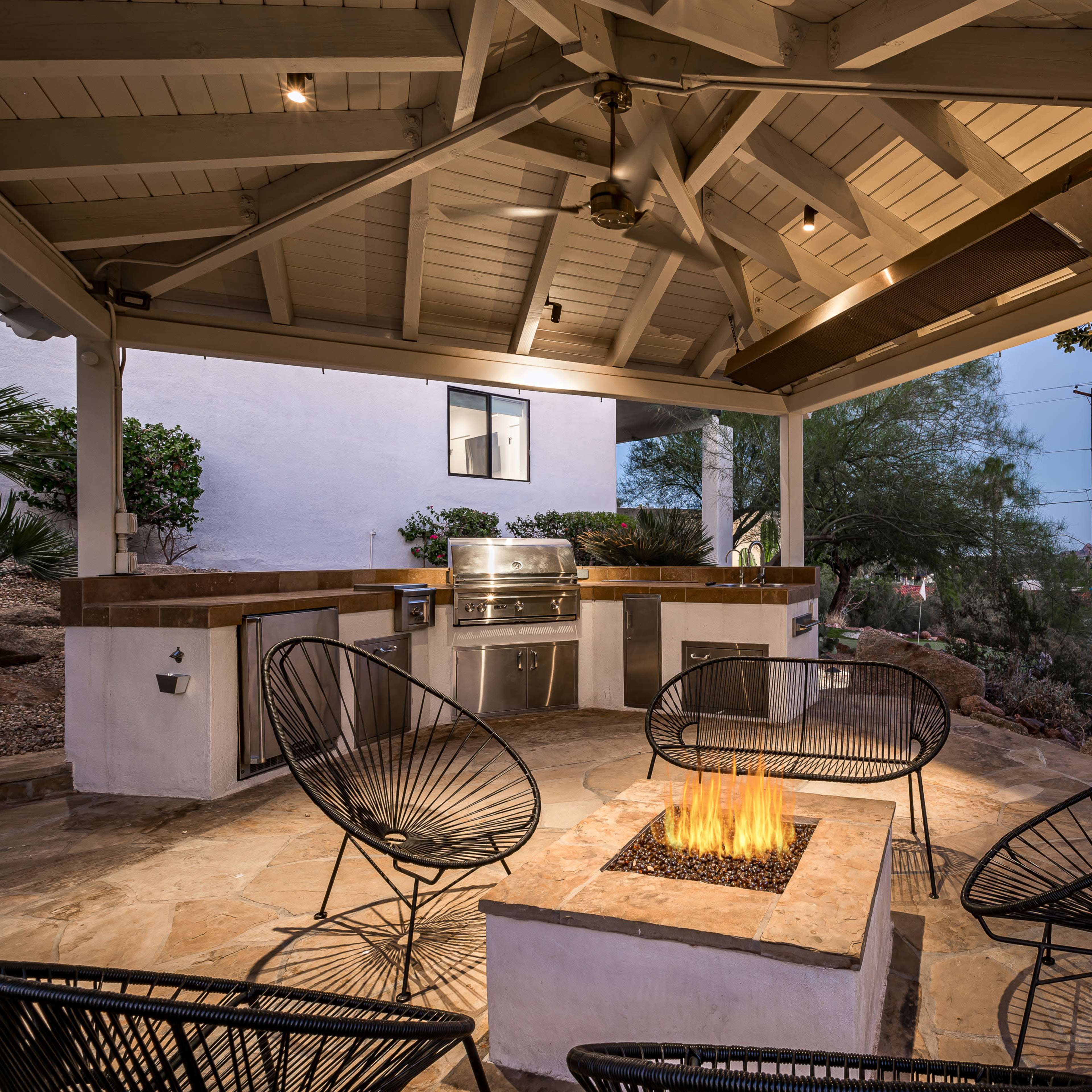 An outdoor patio features a fire pit surrounded by stylish chairs and an adjacent built-in grill area under a covered roof.