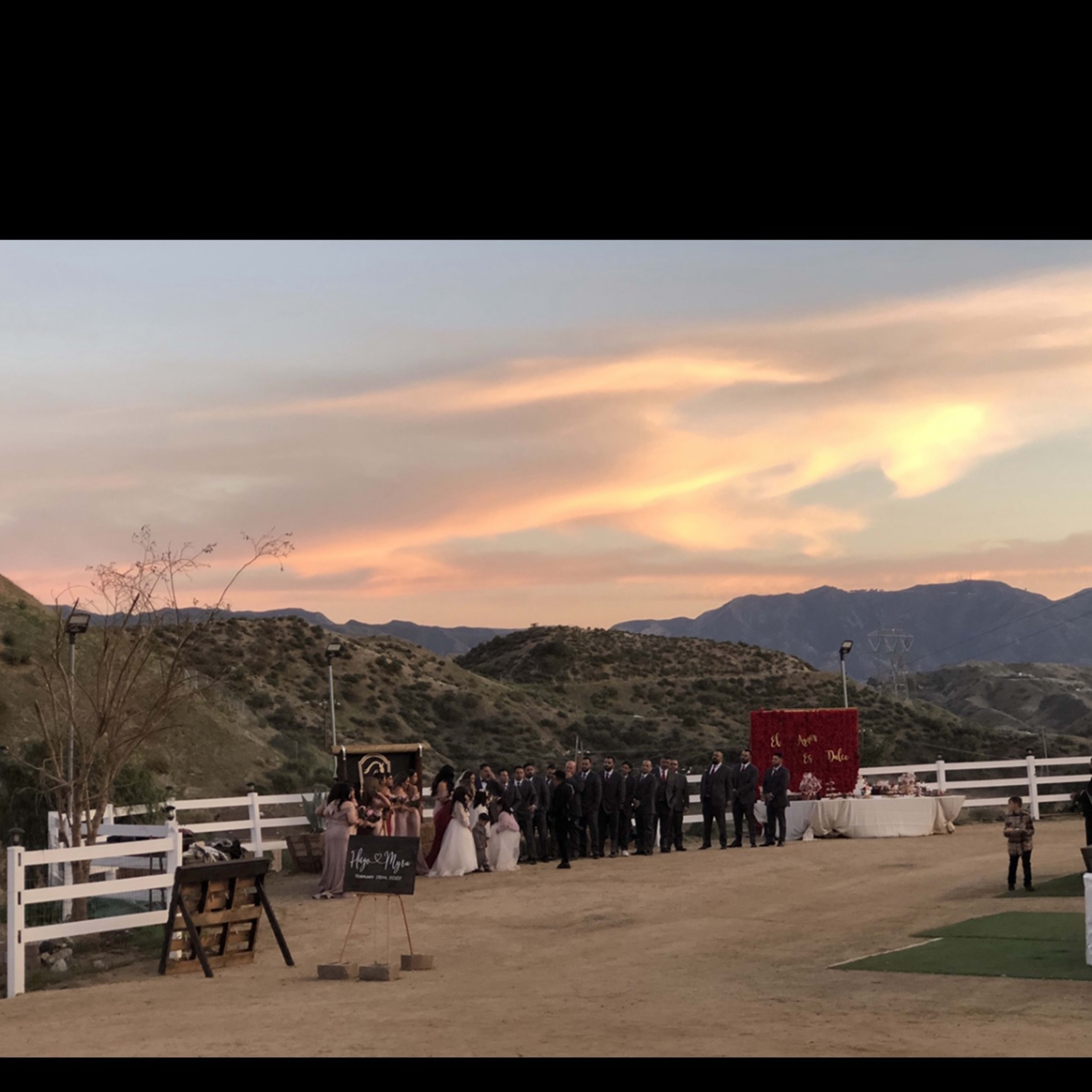 A wedding ceremony is taking place outdoors with a group of people in formal attire standing in front of a bride and groom, against a backdrop of mountains and a colorful sunset.