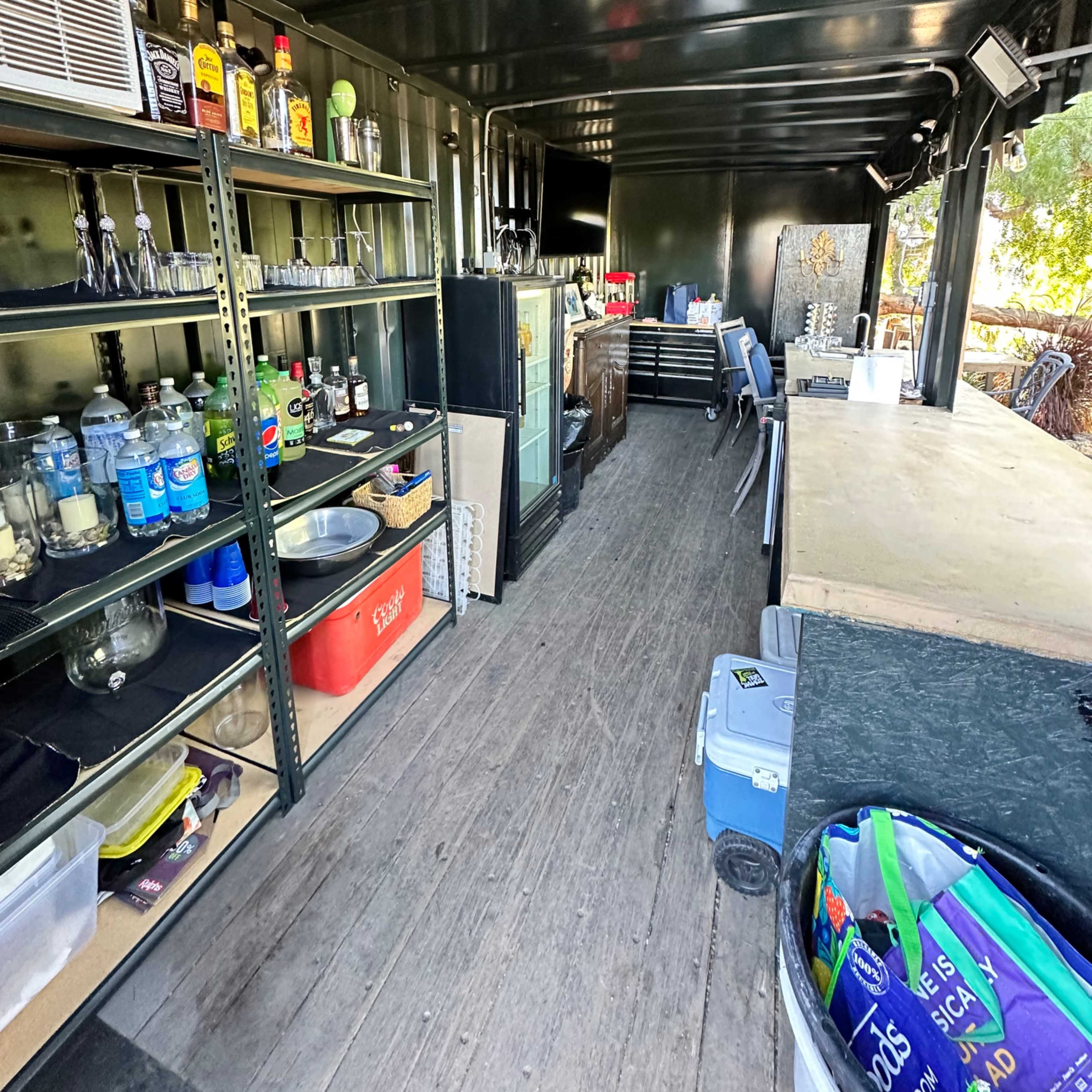 The interior of a commercial kitchen or bar setup inside a shipping container, featuring shelves stocked with beverages and utensils, a large countertop, and kitchen appliances.