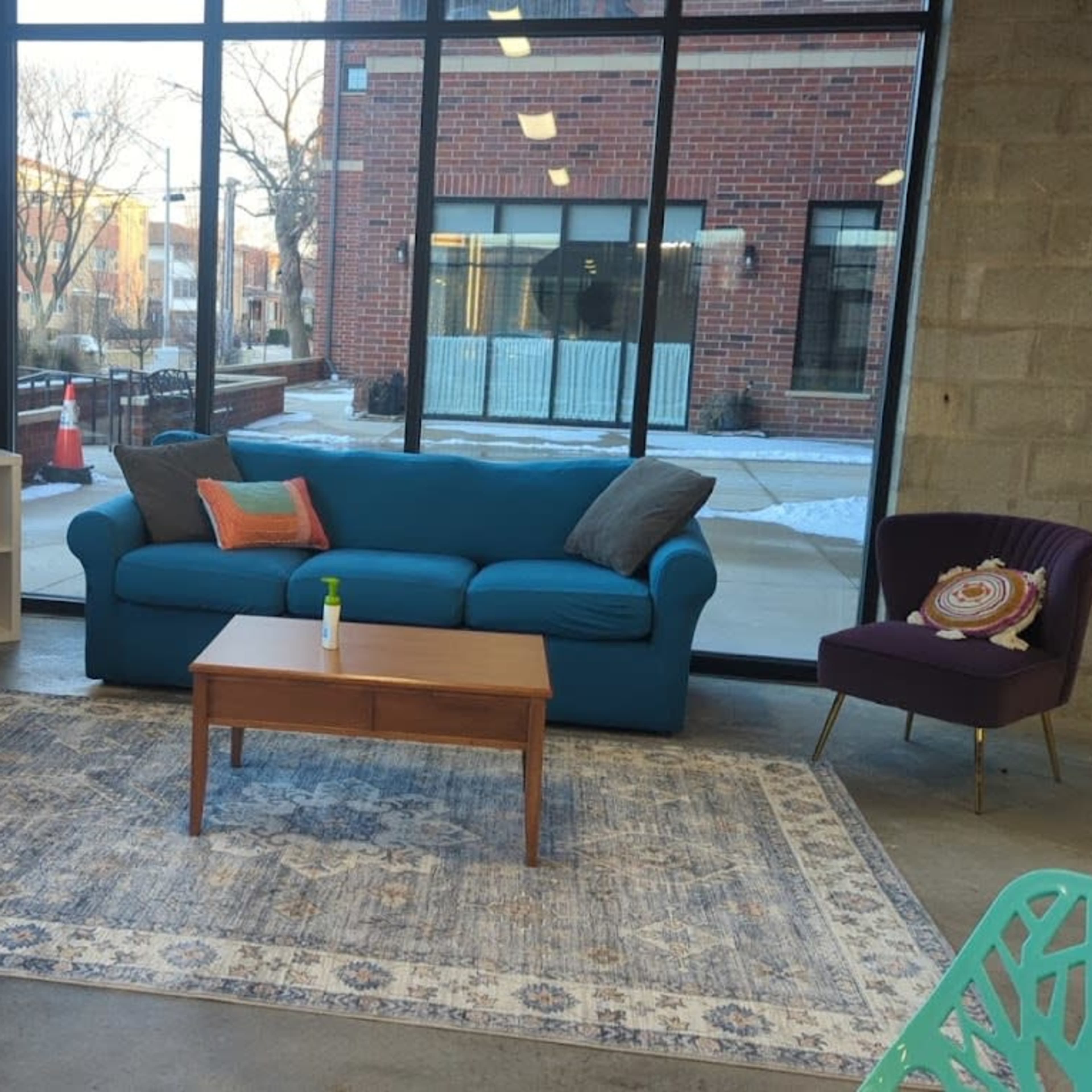 A modern lounge area features two blue sofas, a wooden coffee table, and a patterned rug, framed by large windows showcasing the outdoors.