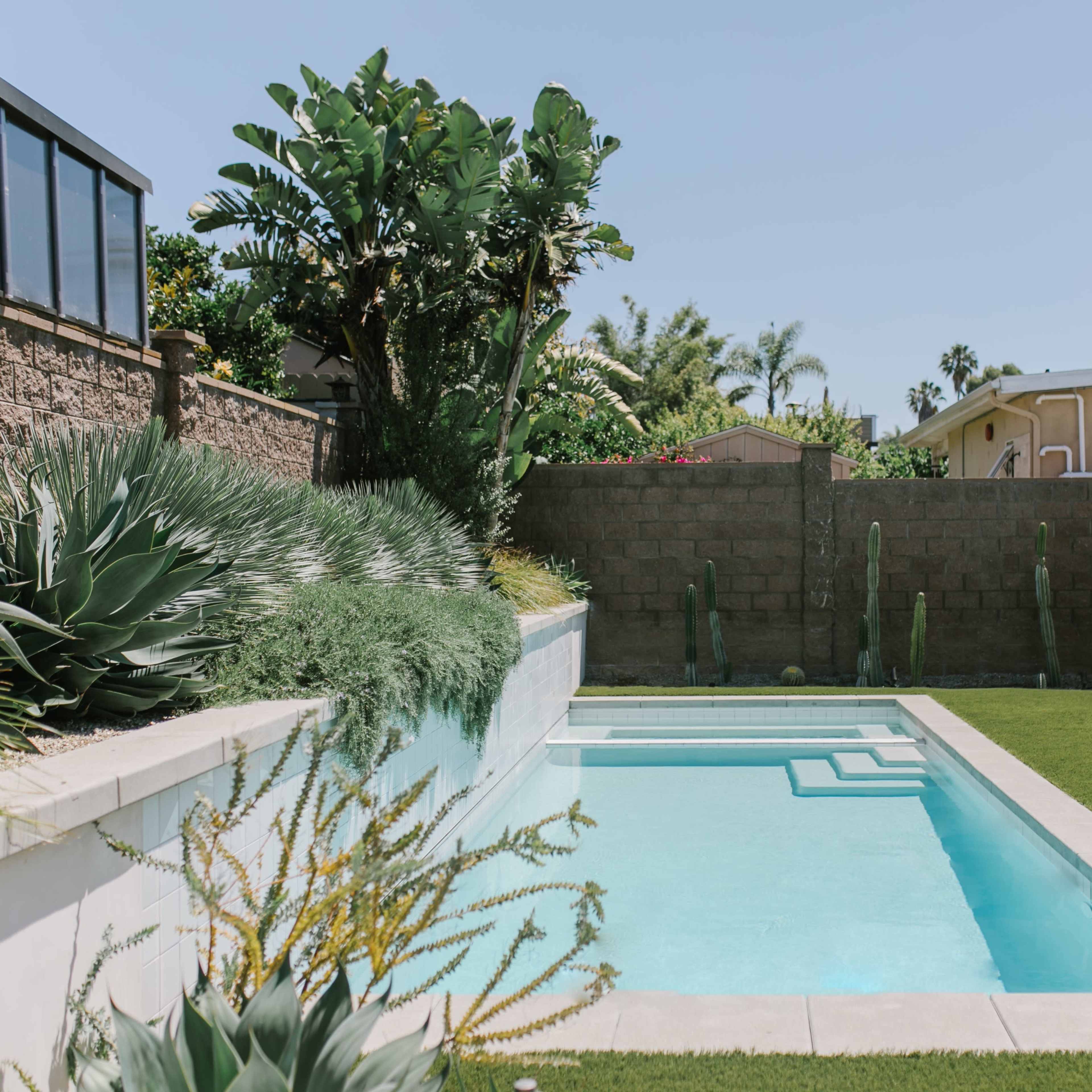 A rectangular swimming pool surrounded by neatly trimmed grass and tropical plants, with two lounge chairs and an umbrella on one side.
