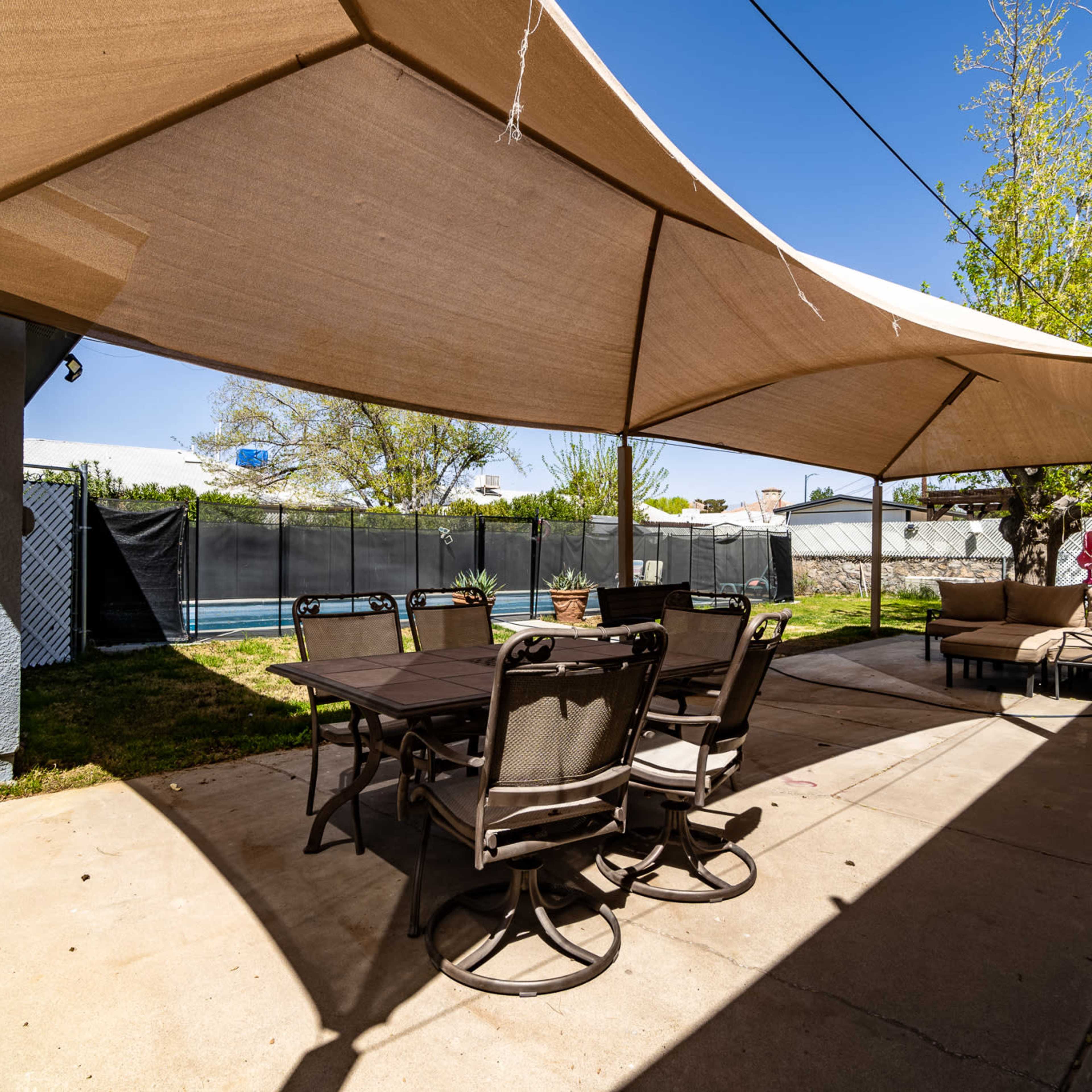 A shaded patio area with a dining table and chairs overlooks a grassy yard surrounded by fencing.