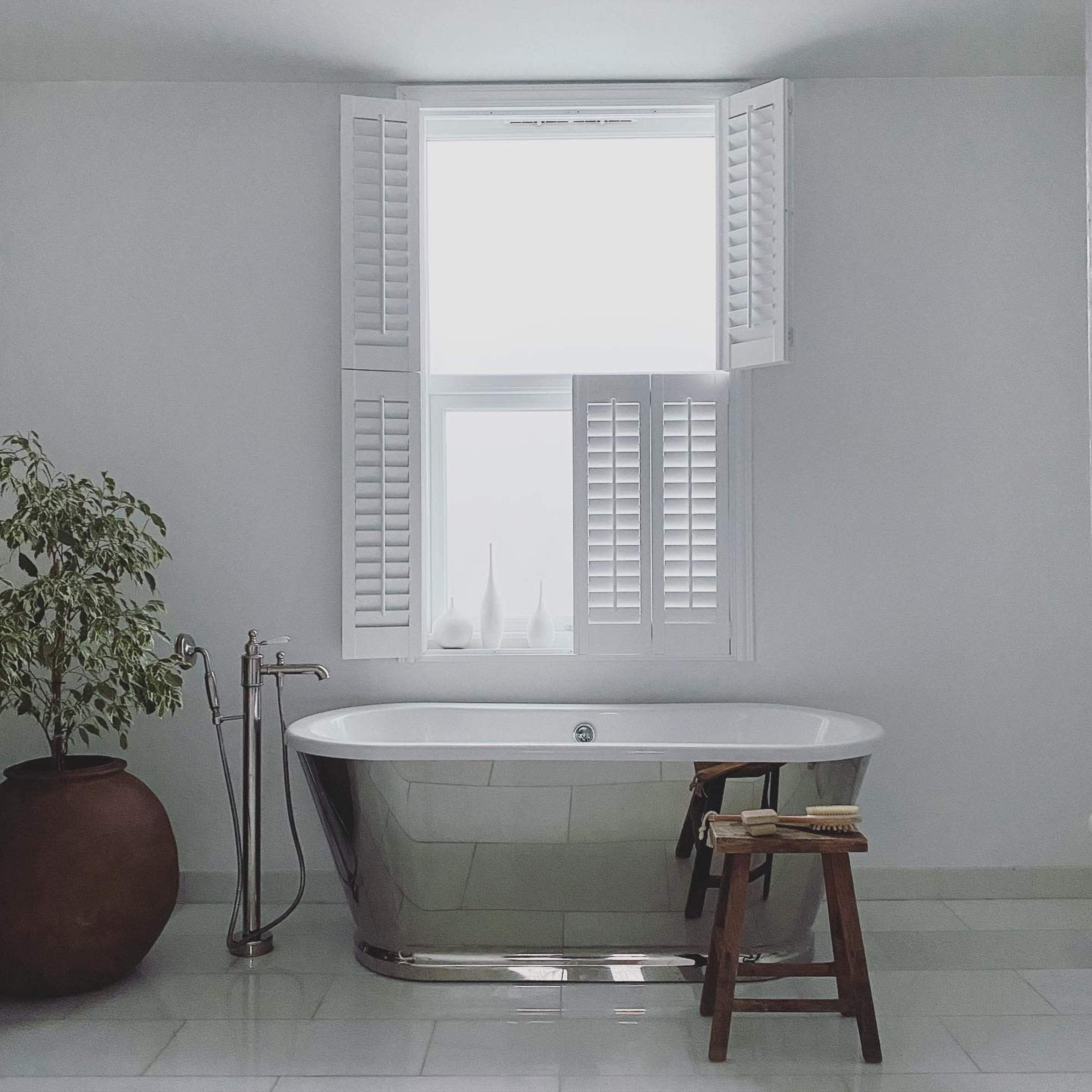 A modern bathroom features a sleek freestanding bathtub beside a potted plant and a wooden stool under a window with louvered shutters.